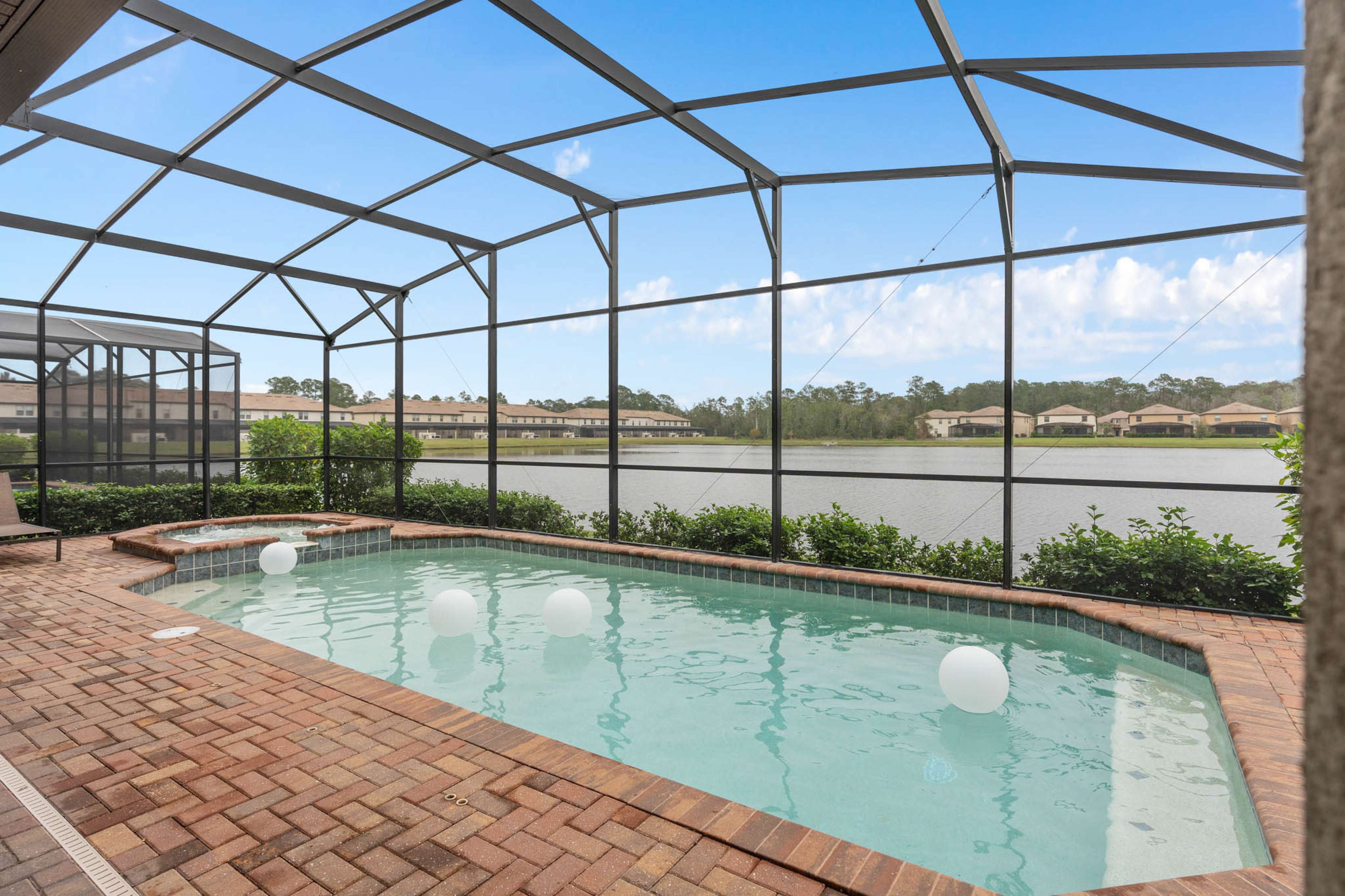 A screened-in pool area with a small spa, surrounded by brick paving and overlooking a calm lake.