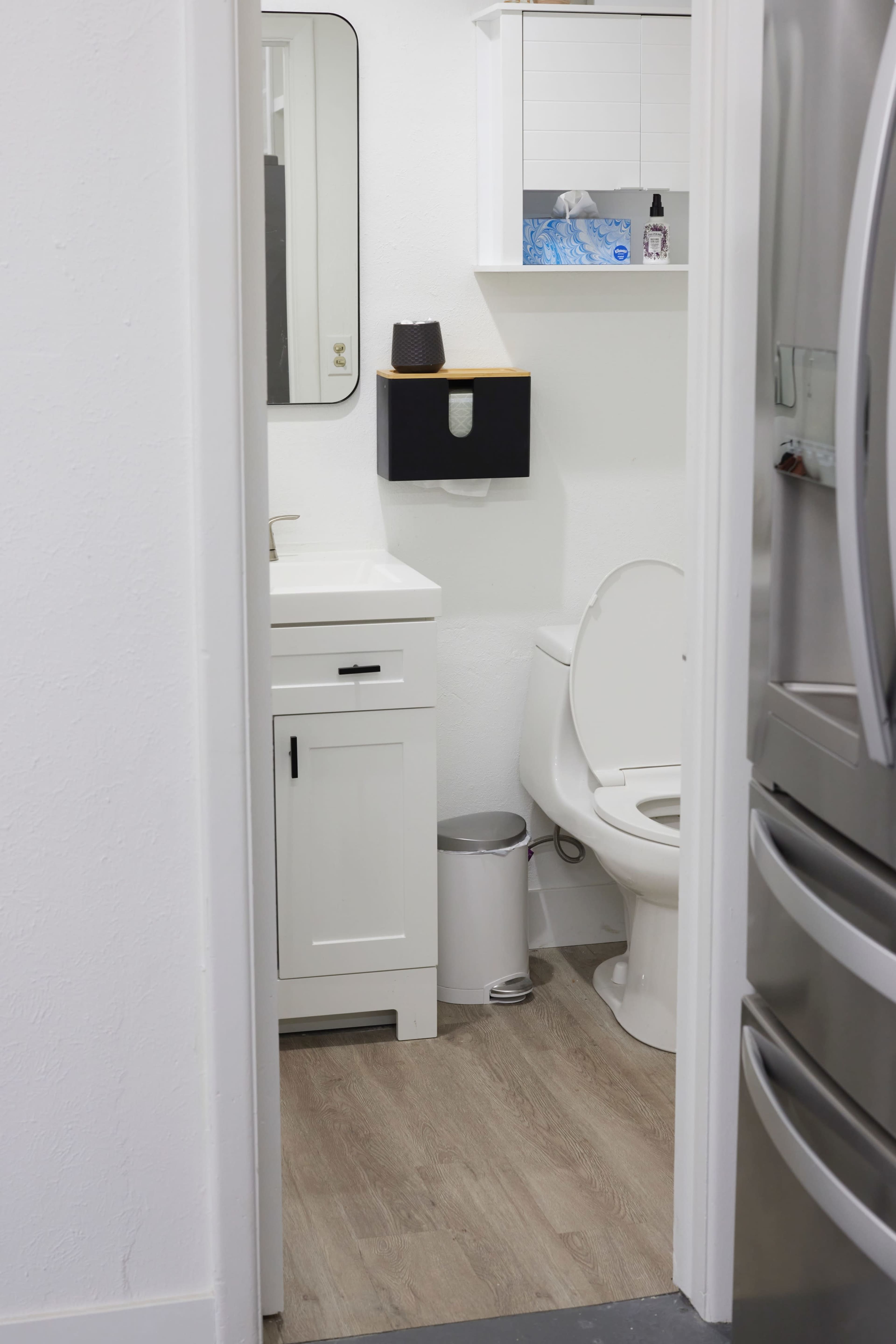 The image shows a small bathroom view from a hallway, featuring a white vanity with a mirror, a toilet, and a wall-mounted shelf.