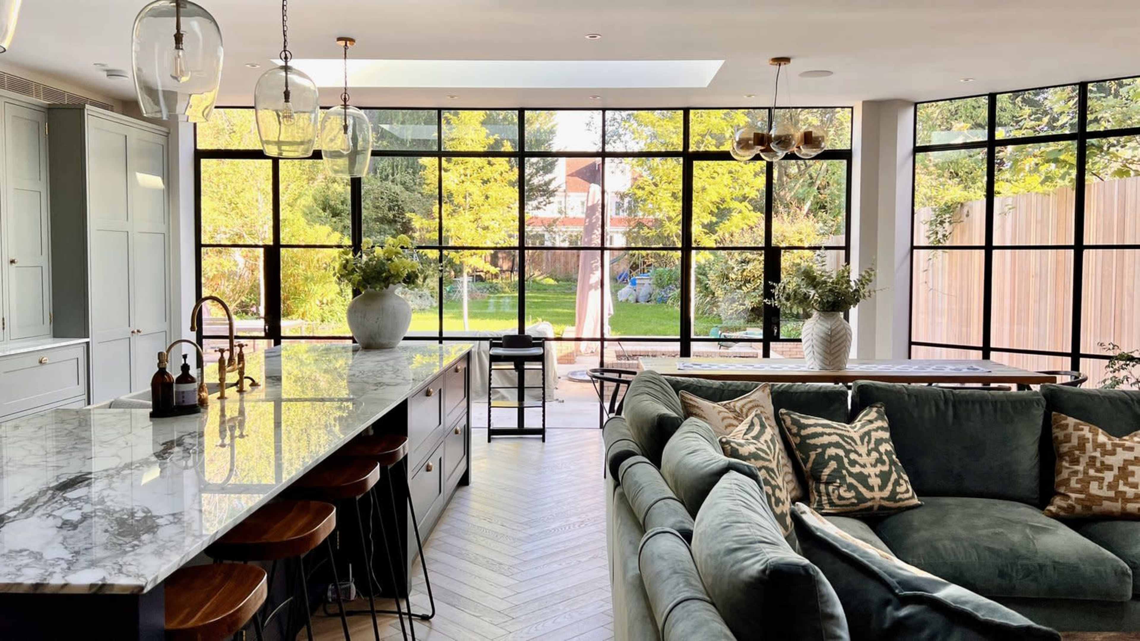 A modern kitchen and living area featuring a marble countertop, a large sectional sofa, and expansive windows overlooking a green garden.