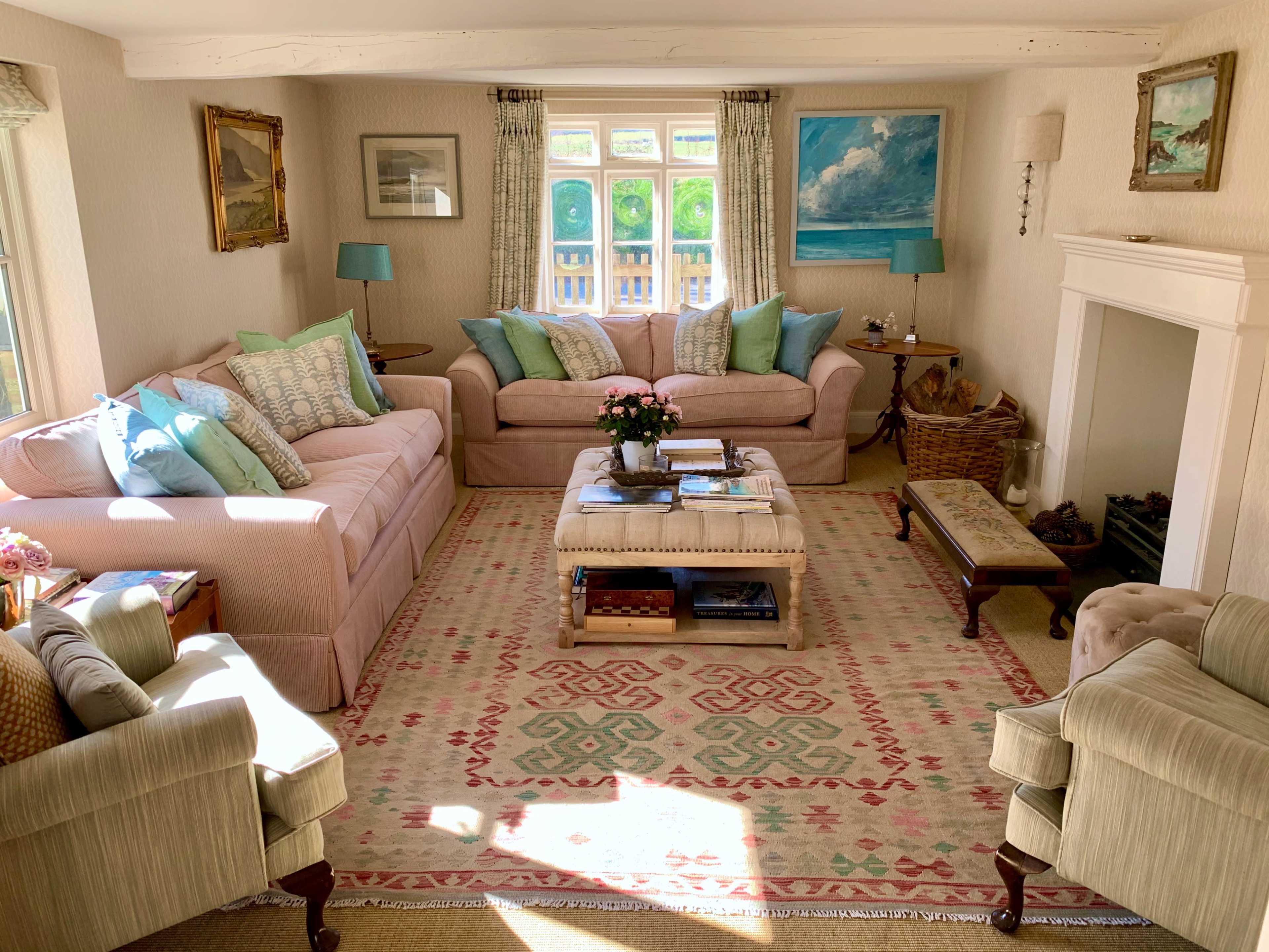 A cozy living room features light pink sofas, decorative pillows, a floral coffee table, and a fireplace, with natural light streaming through the windows.