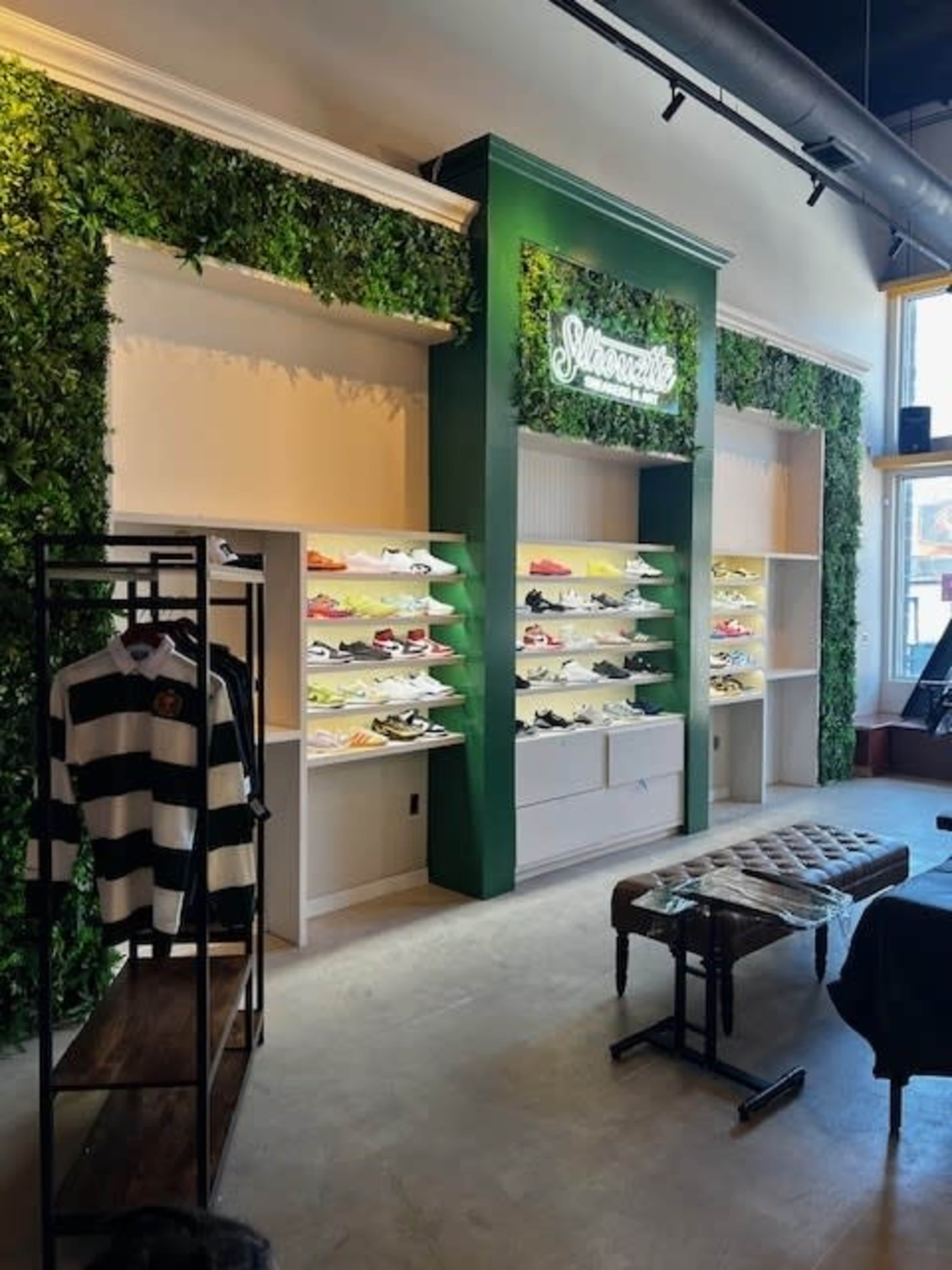 The image shows a modern retail store interior featuring green plant walls, shelving displaying various shoes, and a seating area with a tufted bench.