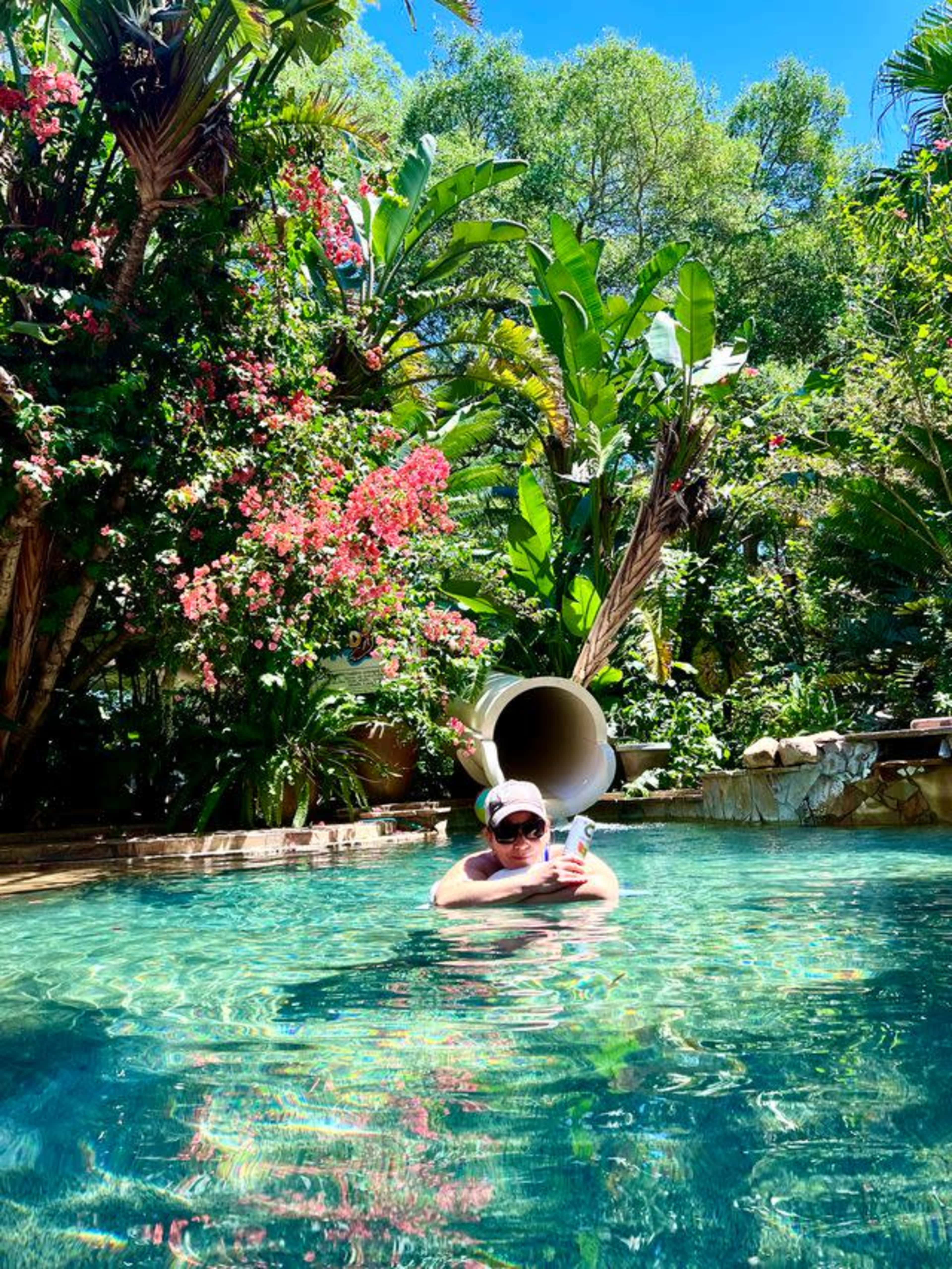 A person relaxes in a clear pool surrounded by lush greenery and vibrant flowers, with a waterslide in the background.