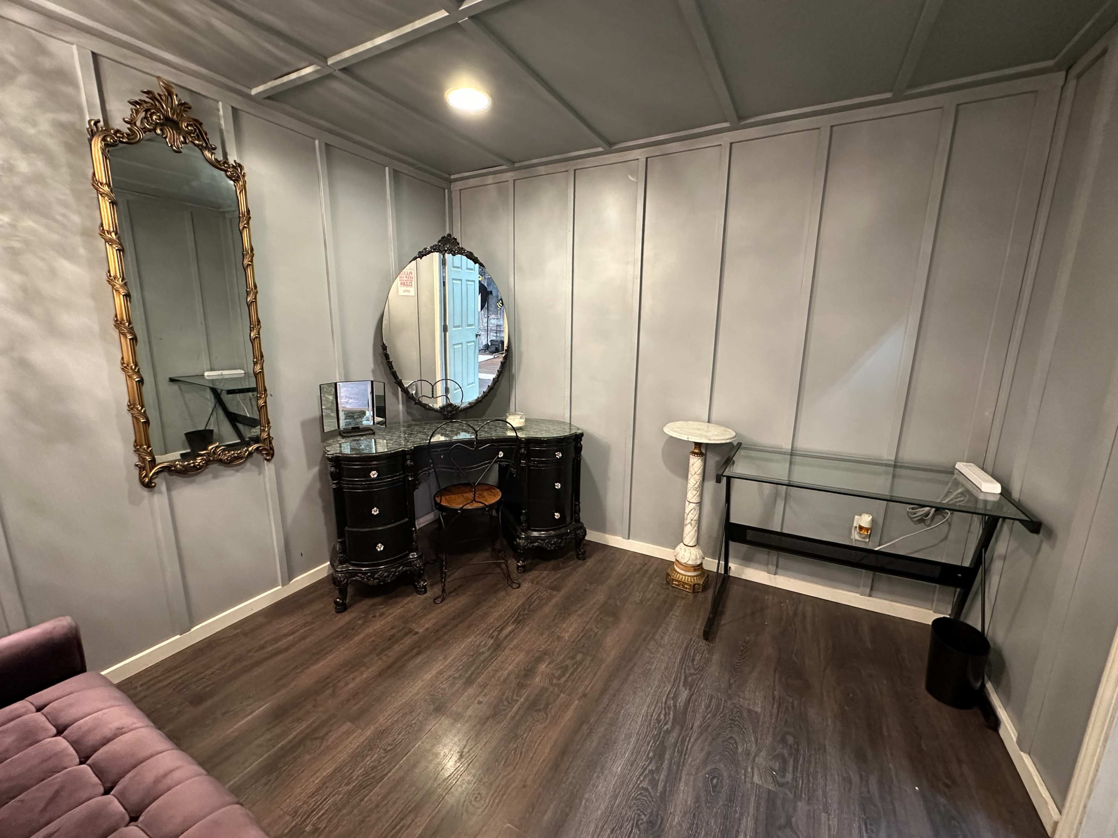 The image shows a small, modern room with gray paneled walls, a velvet couch, a black vanity with a mirror, and a glass table against one wall.