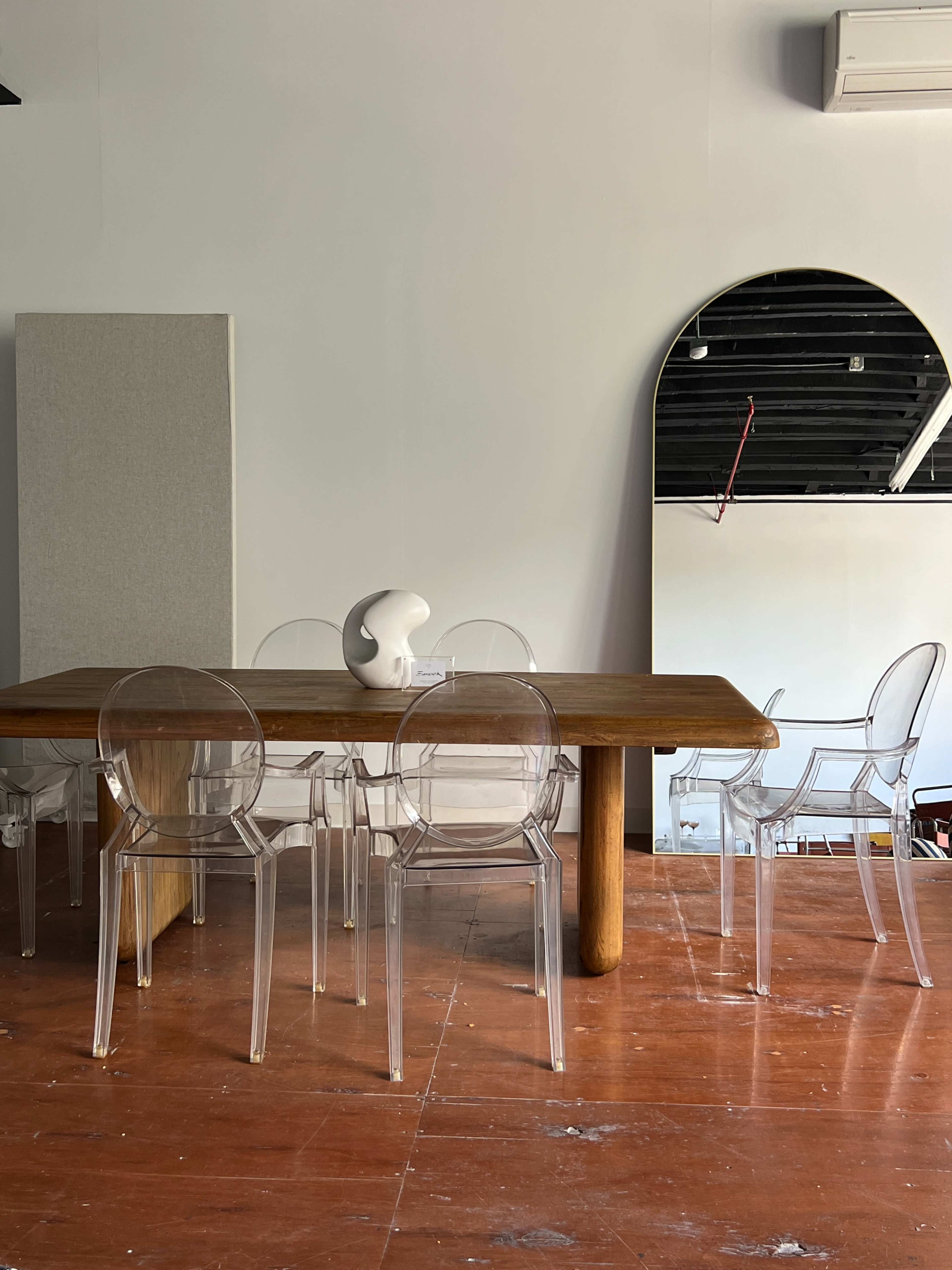 A wooden dining table is surrounded by clear acrylic chairs in a minimalist room with white walls and a large mirror.