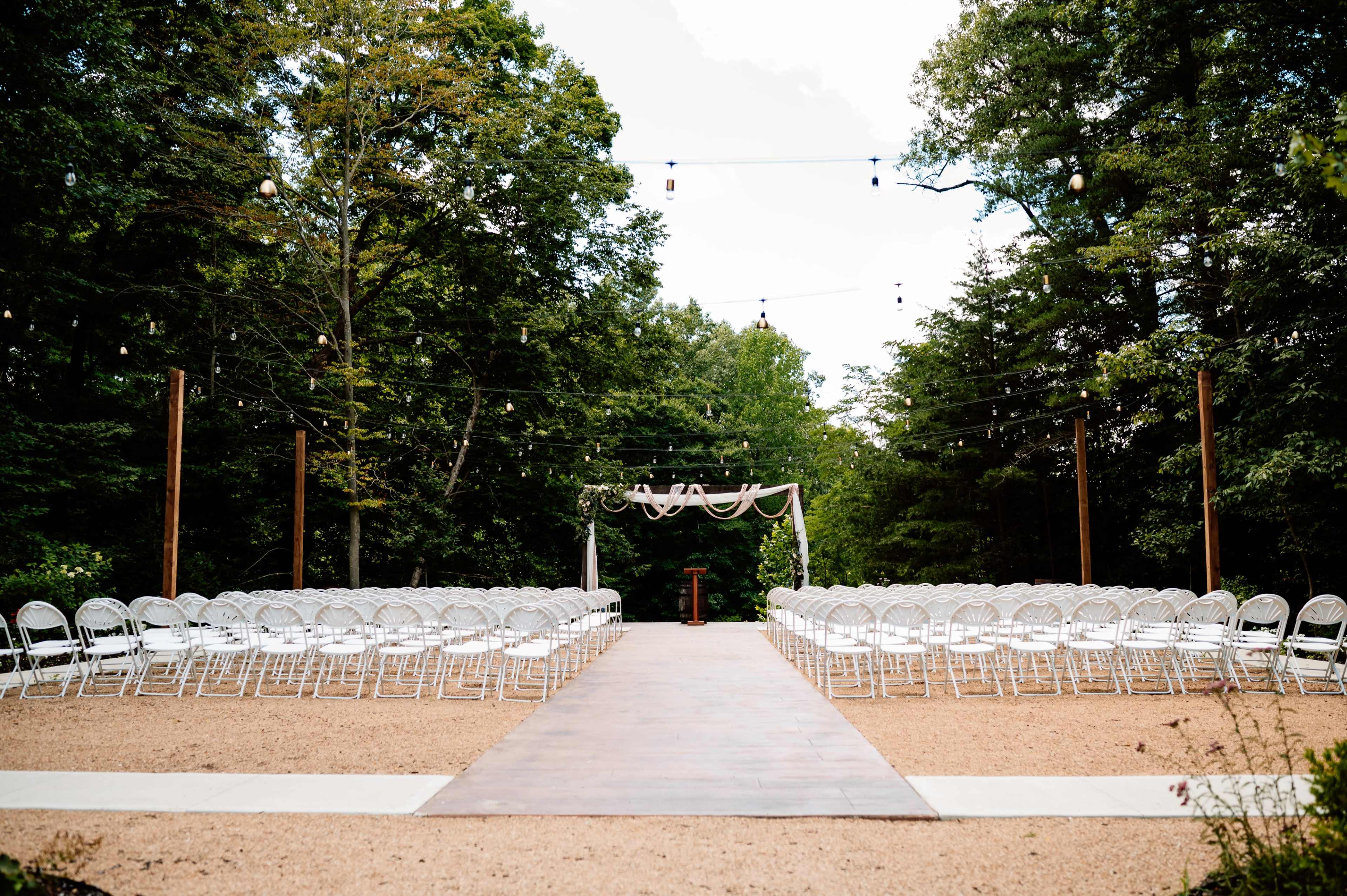 The image shows an outdoor wedding ceremony setup with rows of white chairs arranged on a gravel path leading to an arch adorned with draped fabric and hanging lights in a forested area.