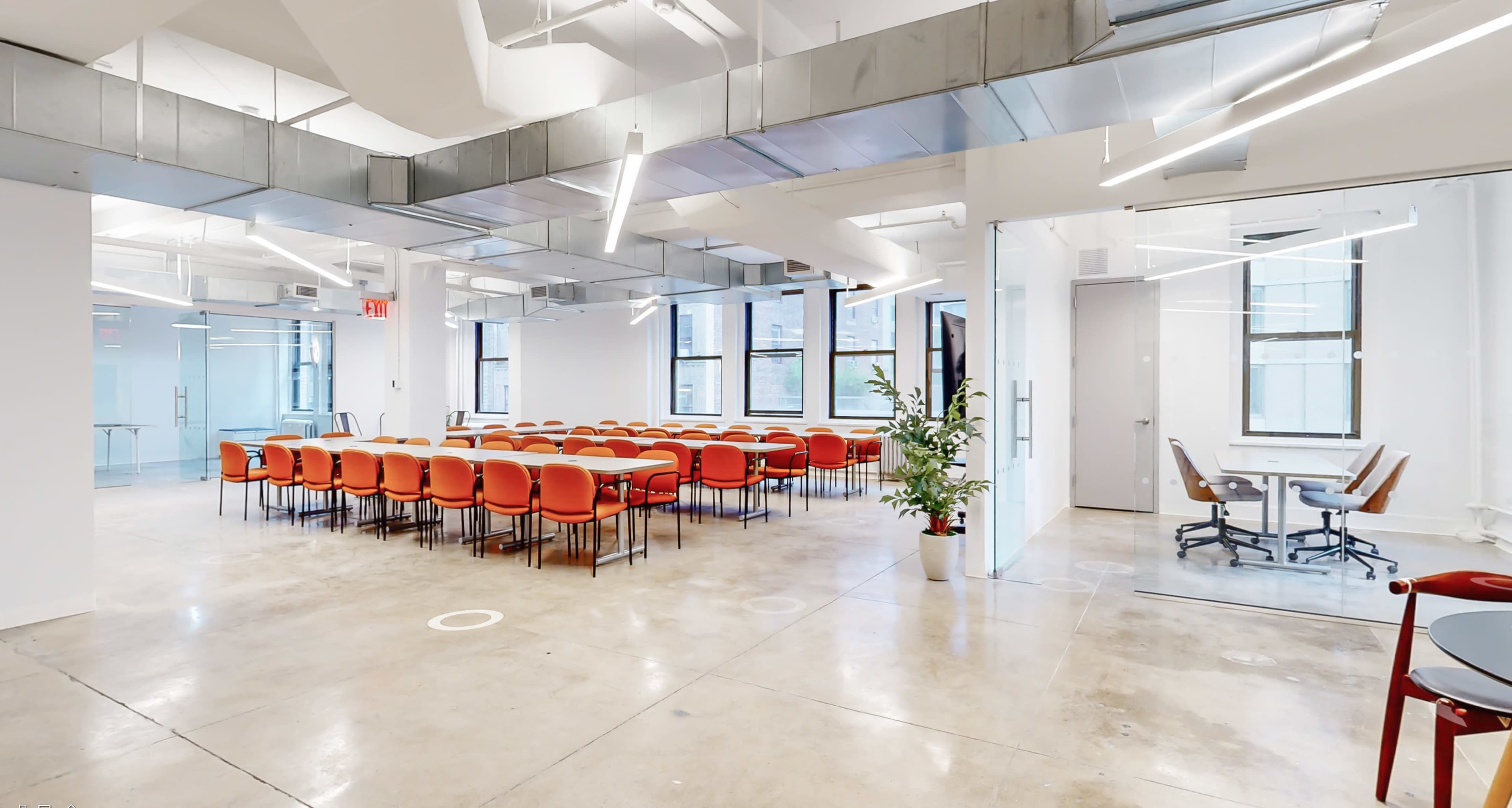 A modern, open space with a large arrangement of orange chairs and tables in the center, surrounded by white walls and bright lighting.