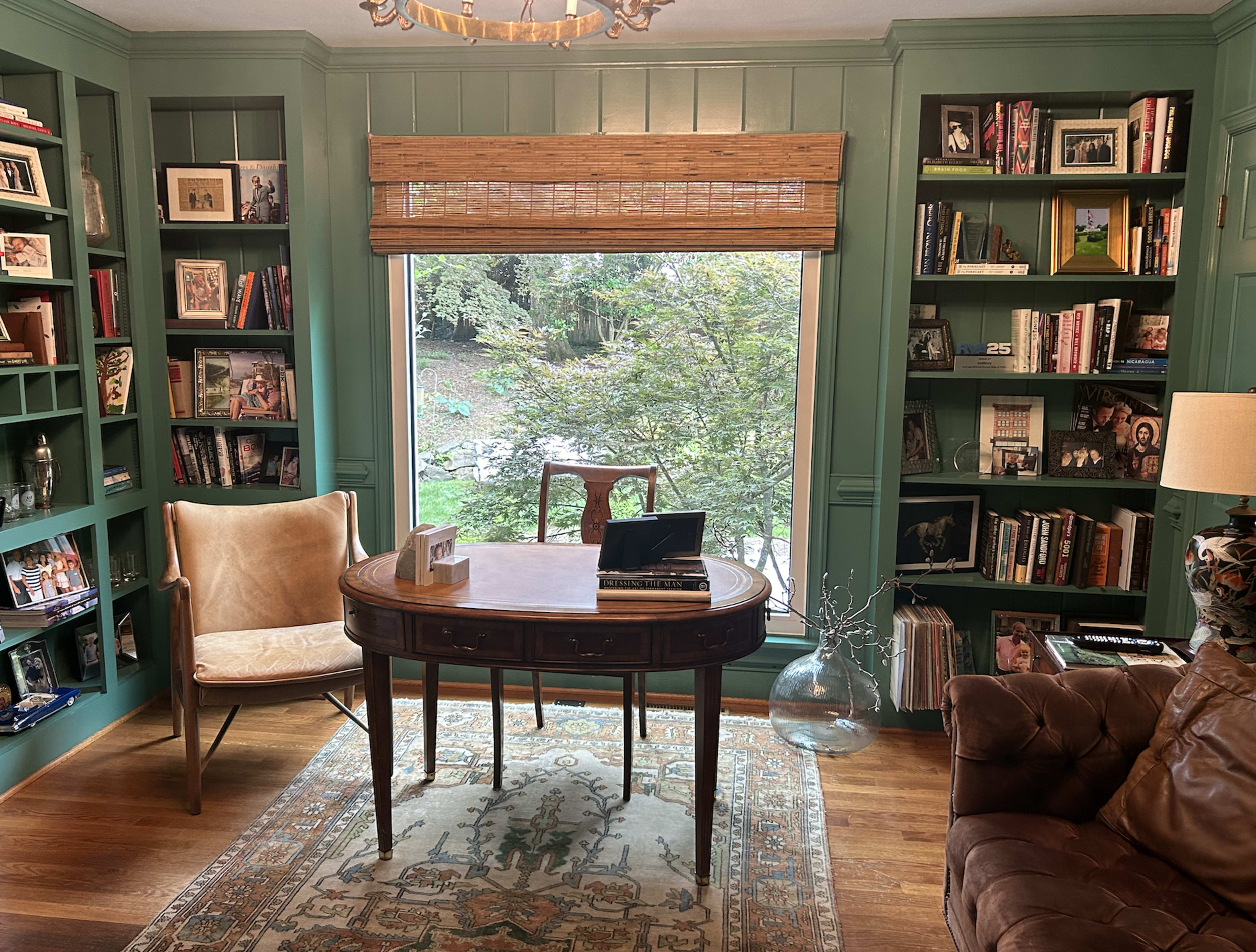 A home office features a wooden desk, a chair, and bookshelves filled with books and photographs, with a large window overlooking a garden.