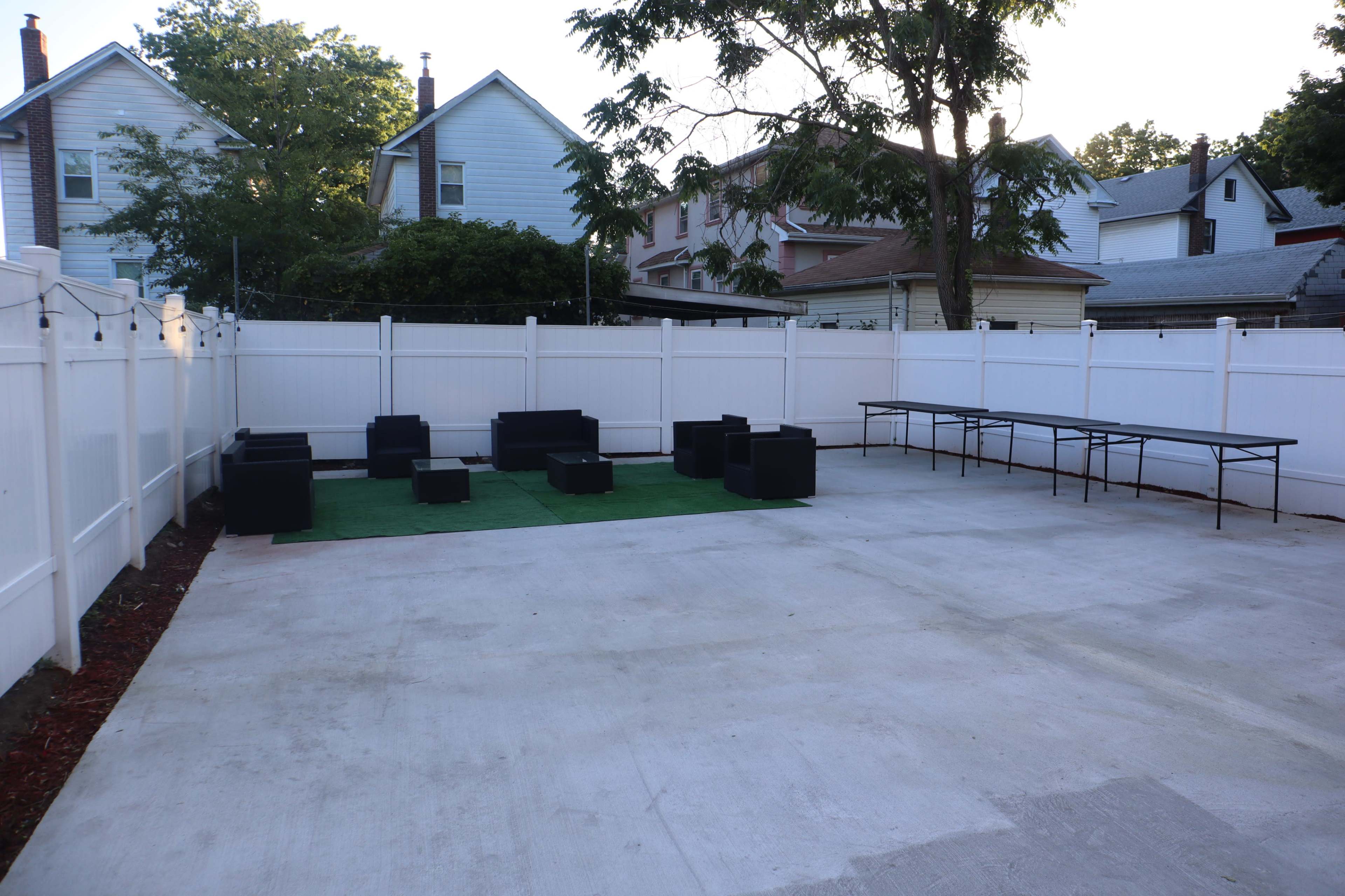 A fenced outdoor area with concrete flooring, featuring a green mat and several black seating arrangements, along with a folding table.