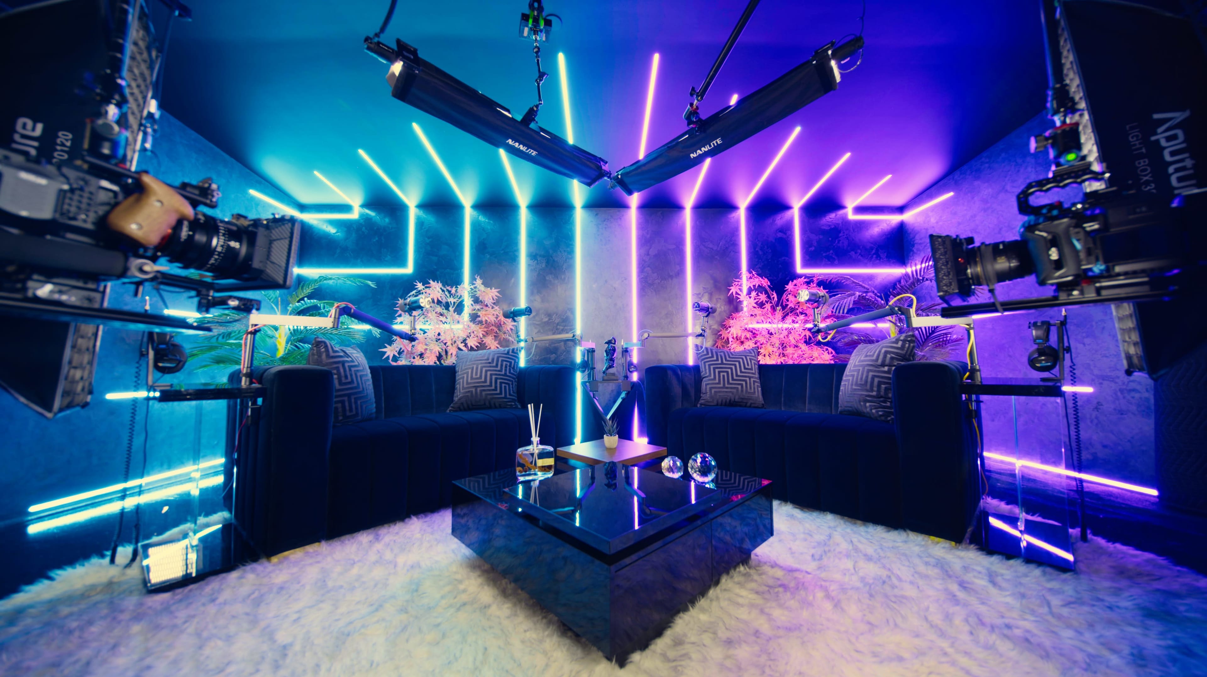 The image shows a modern interior with two black sofas facing each other, illuminated by vibrant neon lights and equipped with cameras on tripods.