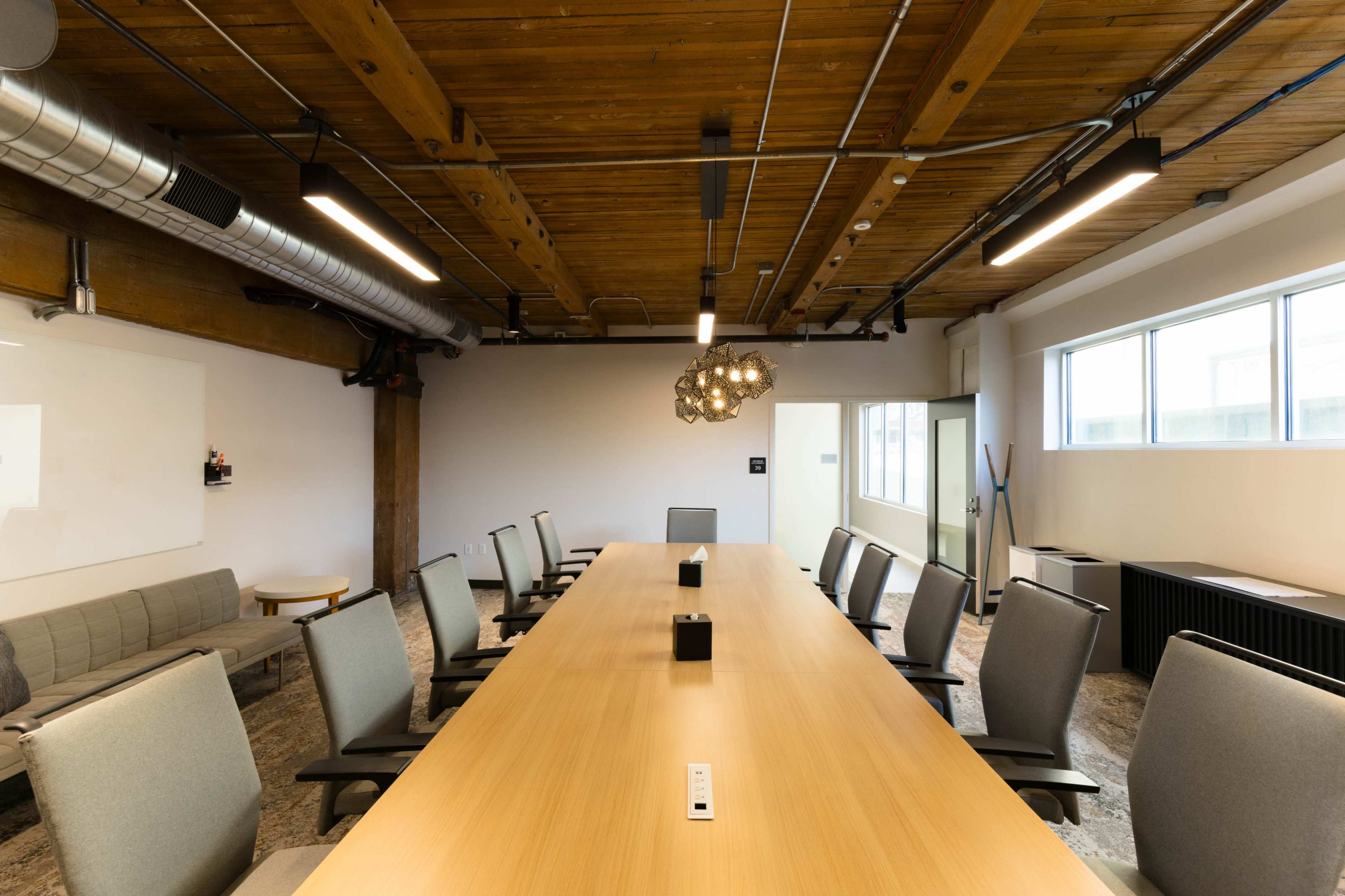 A well-lit conference room features a long wooden table surrounded by ergonomic chairs, with exposed wooden beams and modern lighting.