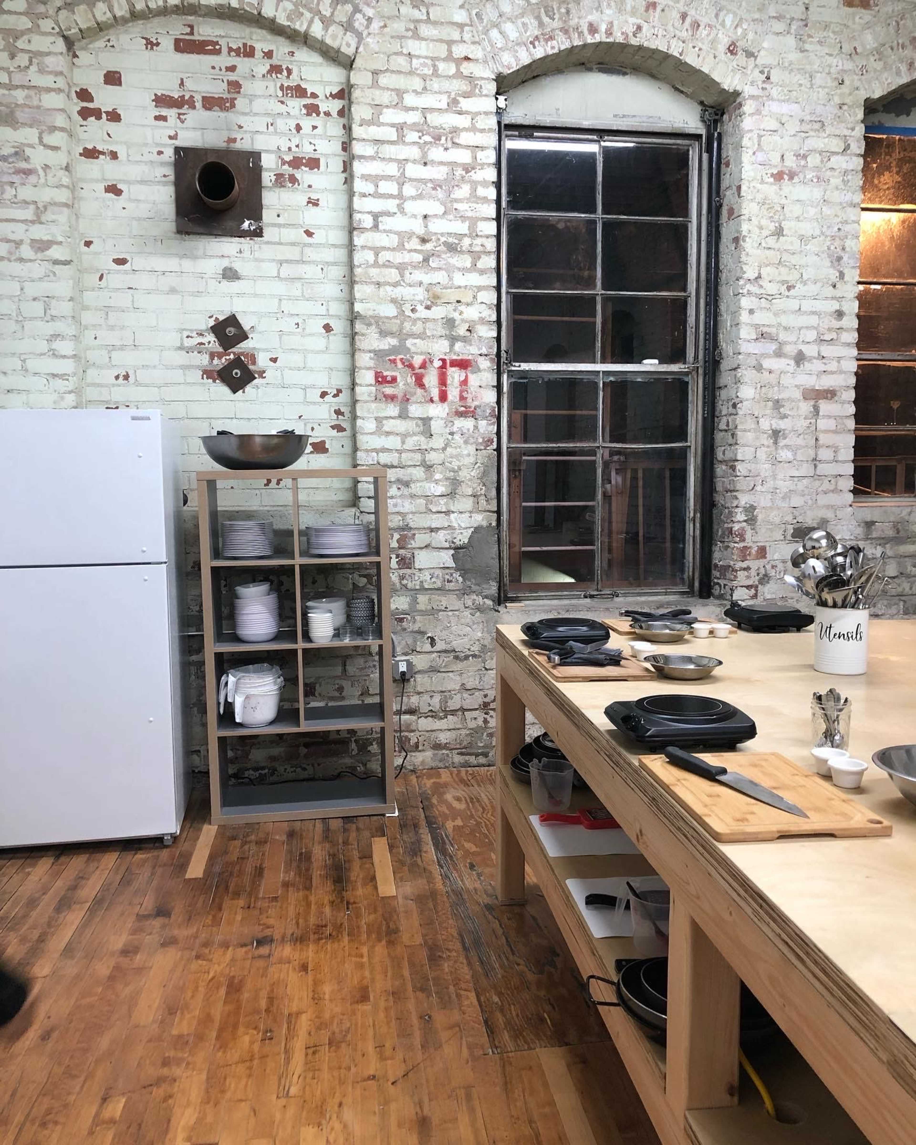 The image shows a rustic kitchen with exposed brick walls, a large wooden table, an open wooden shelf displaying dishware, and a refrigerator against the wall.