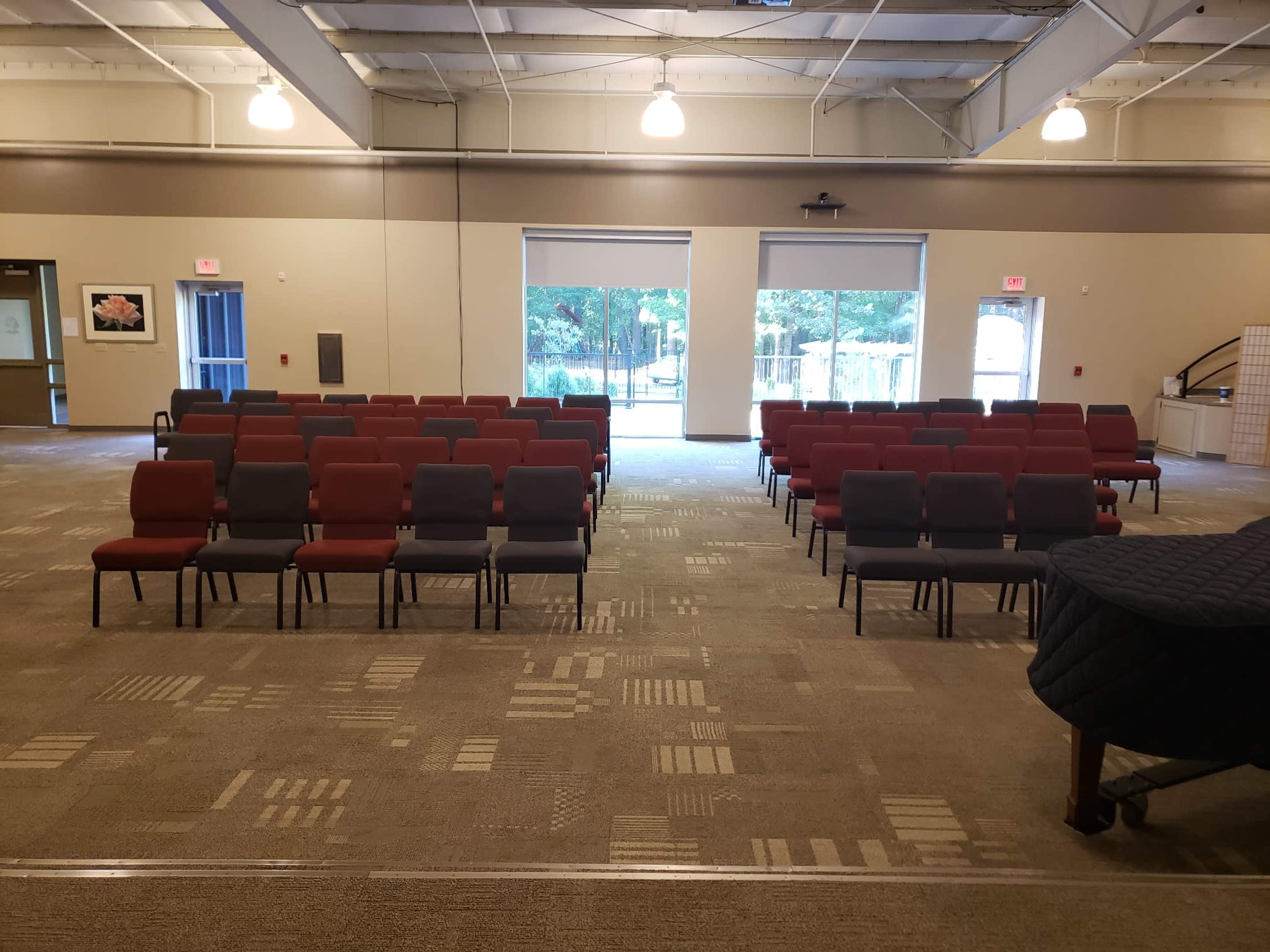 A large room arranged with rows of chairs, alternating between red and gray, facing a window with a view of greenery outside.