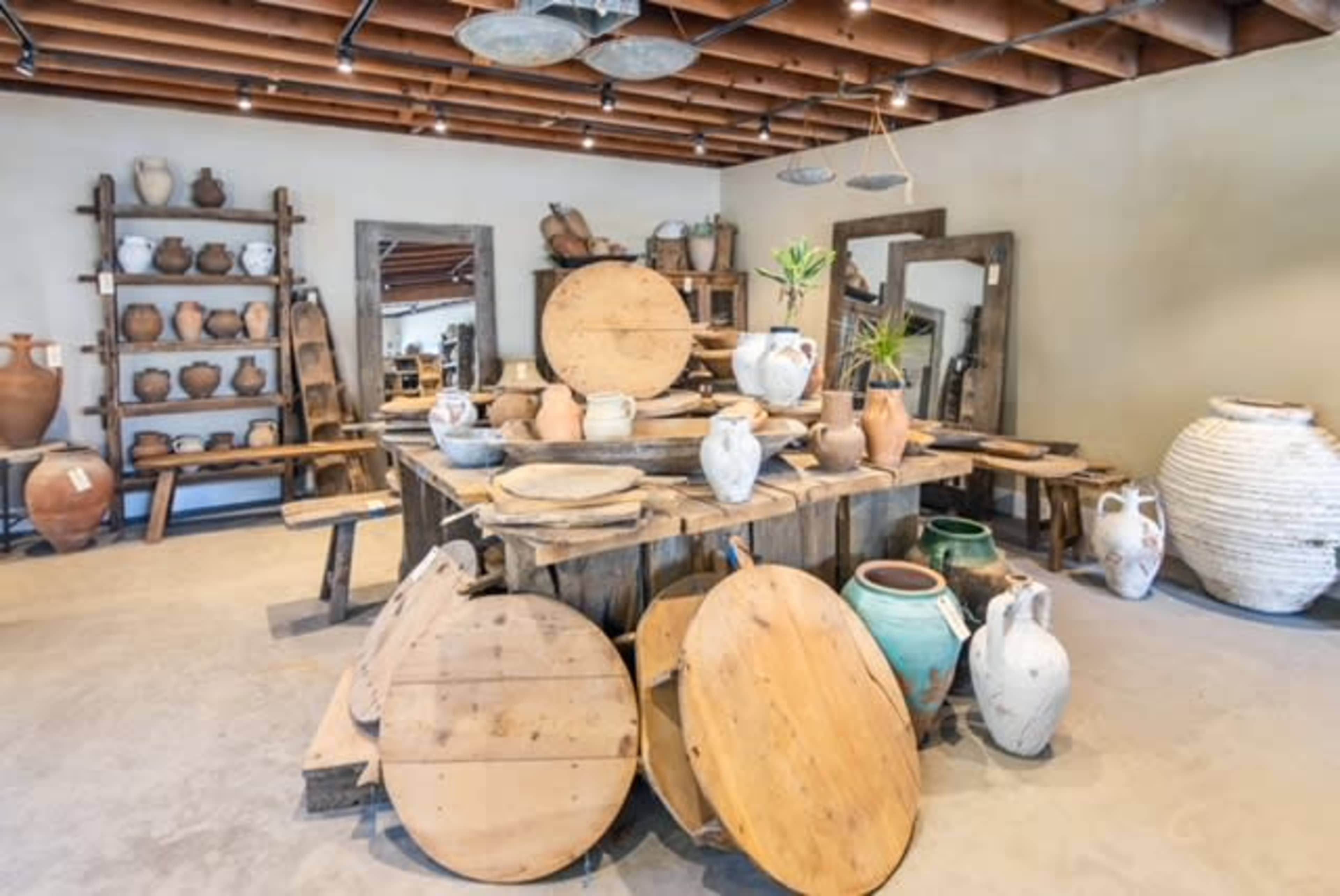 The image shows a rustic shop interior filled with a variety of pottery and wooden furniture arranged around a central wooden table.