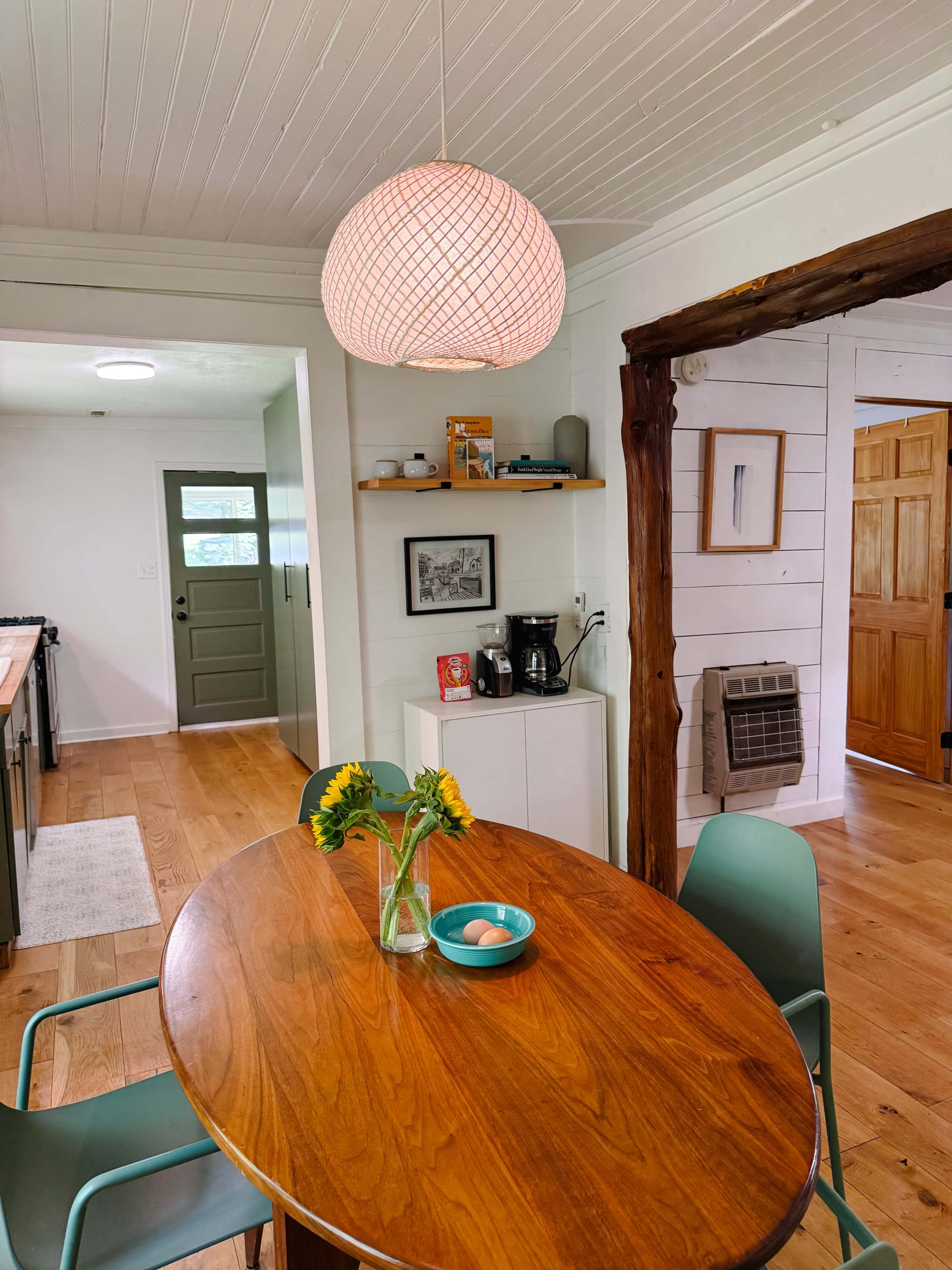 A wooden dining table with a vase of sunflowers and a bowl of eggs is surrounded by green chairs, with a doorway leading to another room in the background.