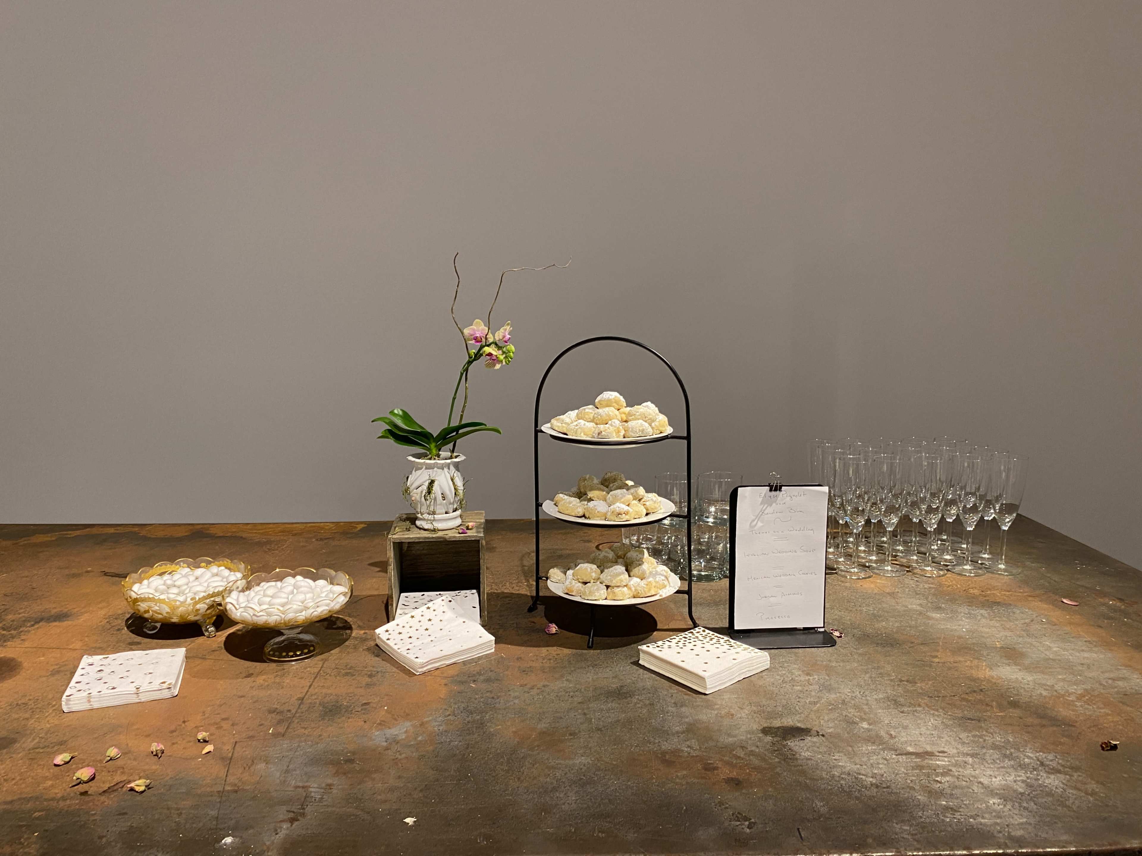 A table displays three tiers of pastries, a small vase with an orchid, a clipboard with a menu, and several empty glasses arranged neatly in the background.