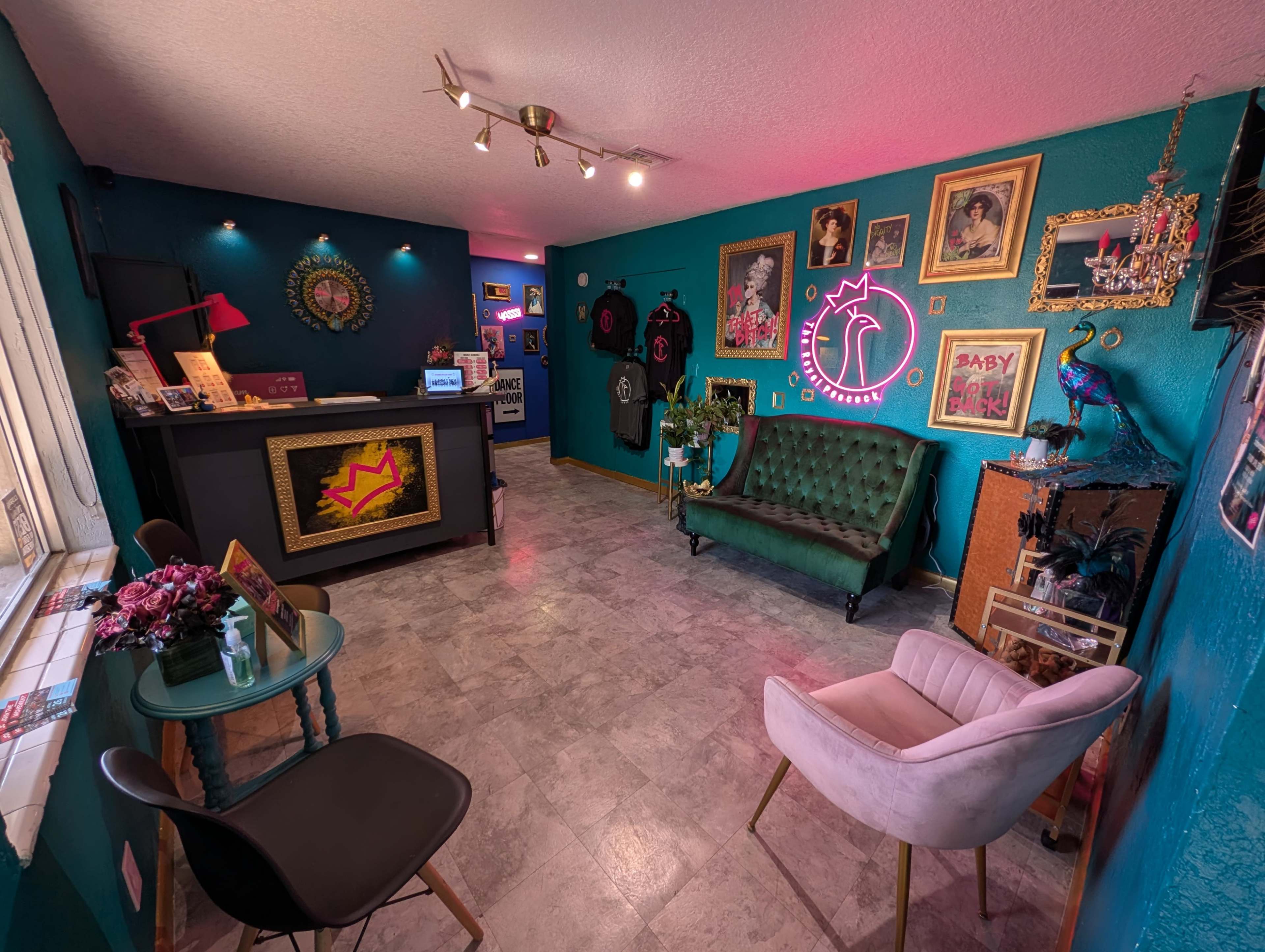 The image shows a colorful tattoo studio featuring a reception desk, green velvet seating, and various art pieces on the walls.