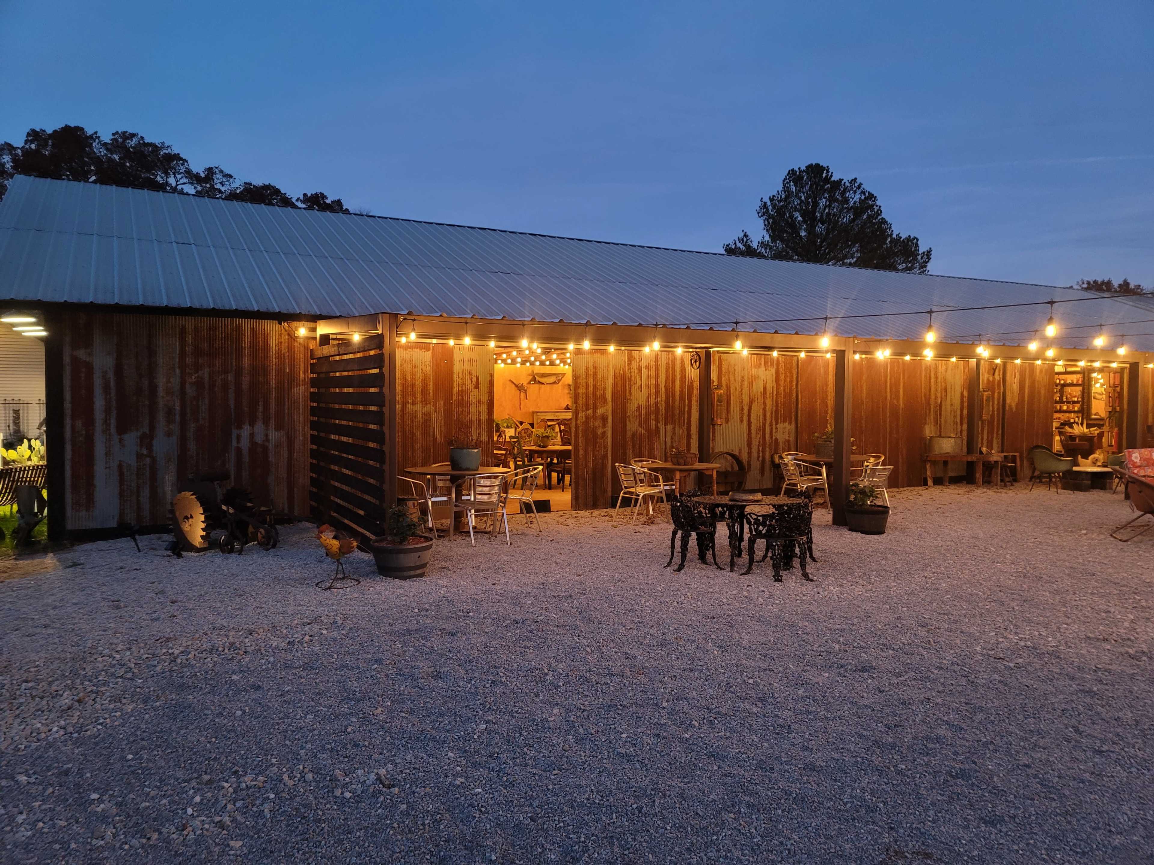 A rustic building with a metal exterior is illuminated by string lights, surrounded by gravel and outdoor seating.