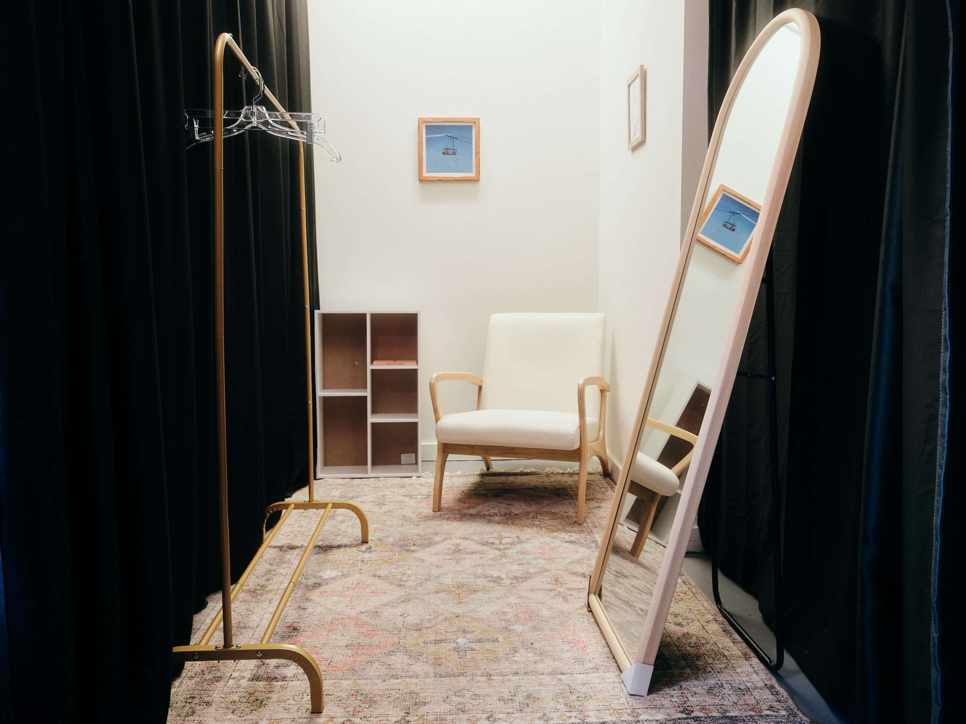 The image shows a small dressing area with a full-length mirror, a clothing rack, a chair, and a decorative wall frame, all set against black curtains.
