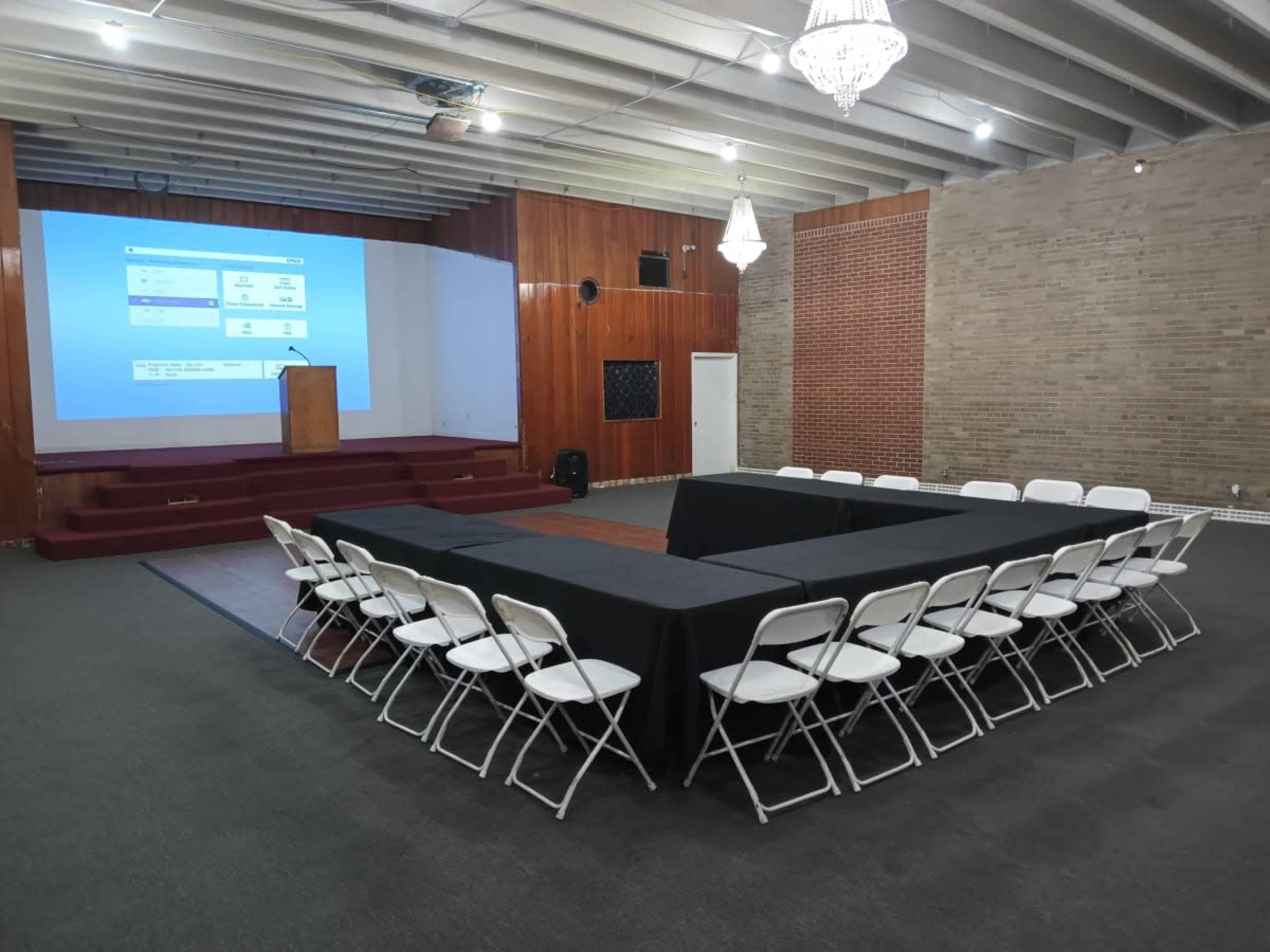 A rectangular setup of tables covered with black cloth and surrounded by white chairs is arranged in a spacious room with a presentation screen and wooden paneling at one end.