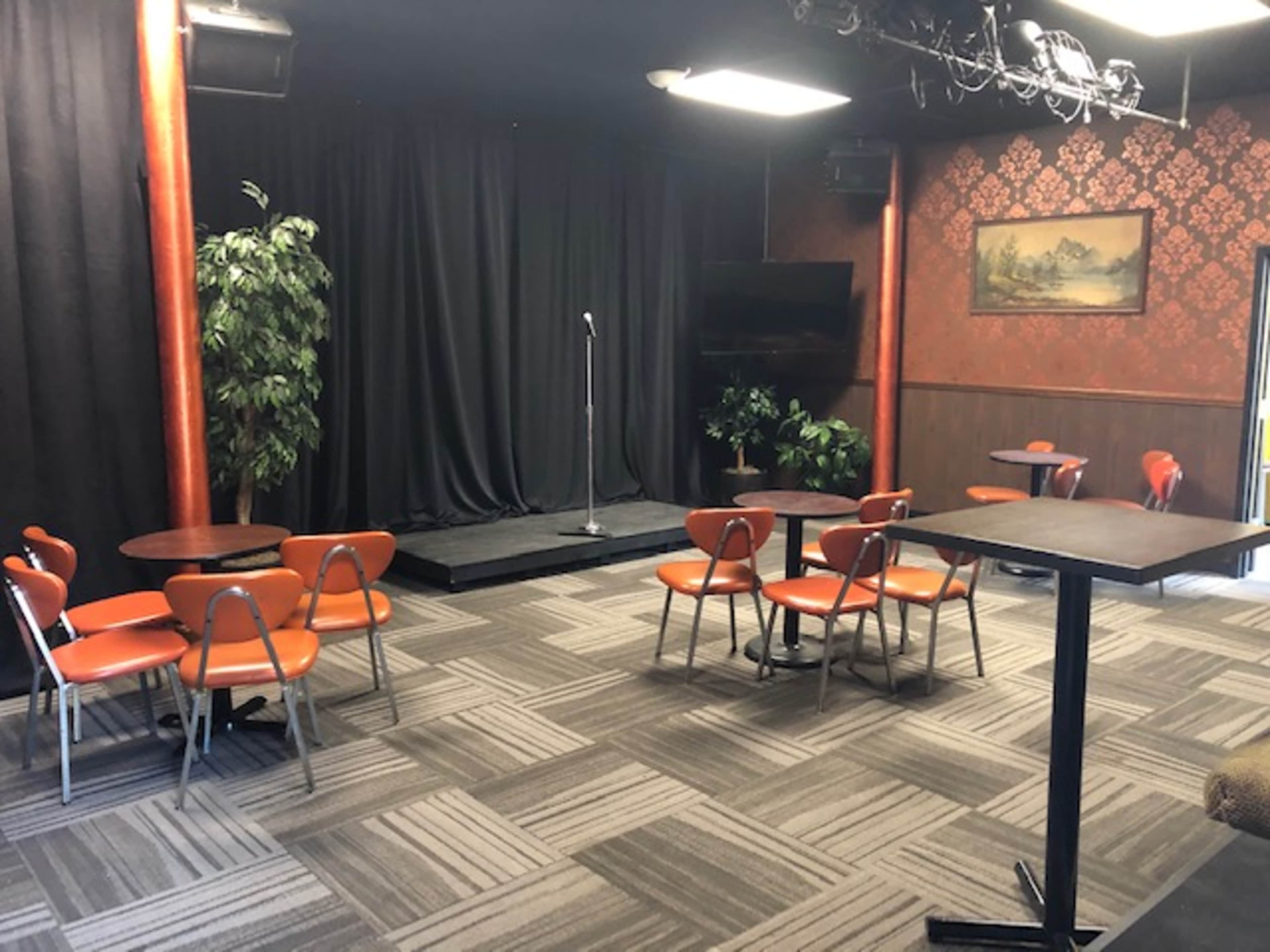 The image shows a small performance stage with a microphone stand, surrounded by several orange chairs and tables in a room with dark walls and a patterned wallpaper.