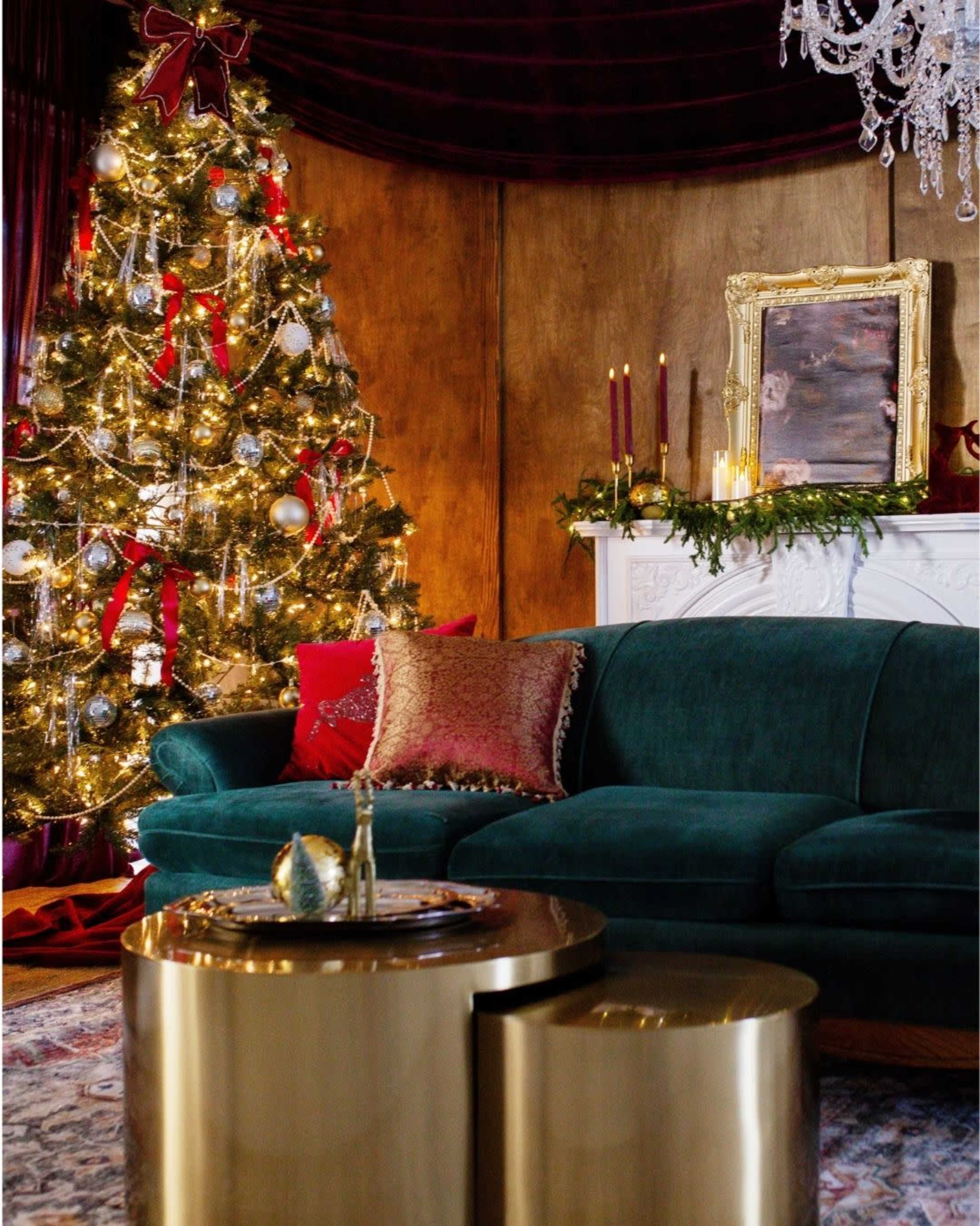 A cozy living room features a decorated Christmas tree, a green couch with decorative pillows, and a gold coffee table in front of a fireplace adorned with candles and a garland.