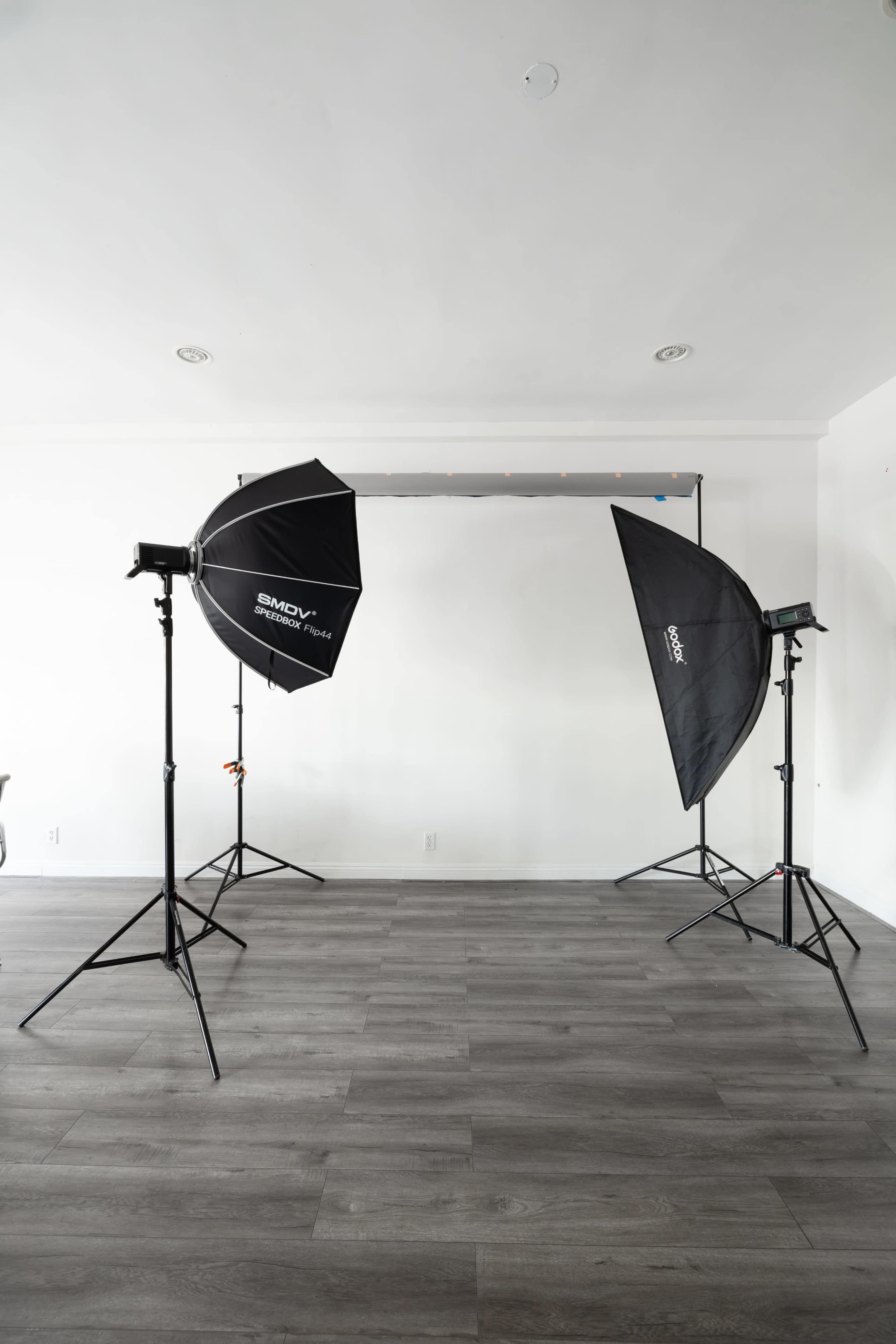 A photography studio setup features two softbox lighting devices on stands facing a backdrop in a minimally furnished room.