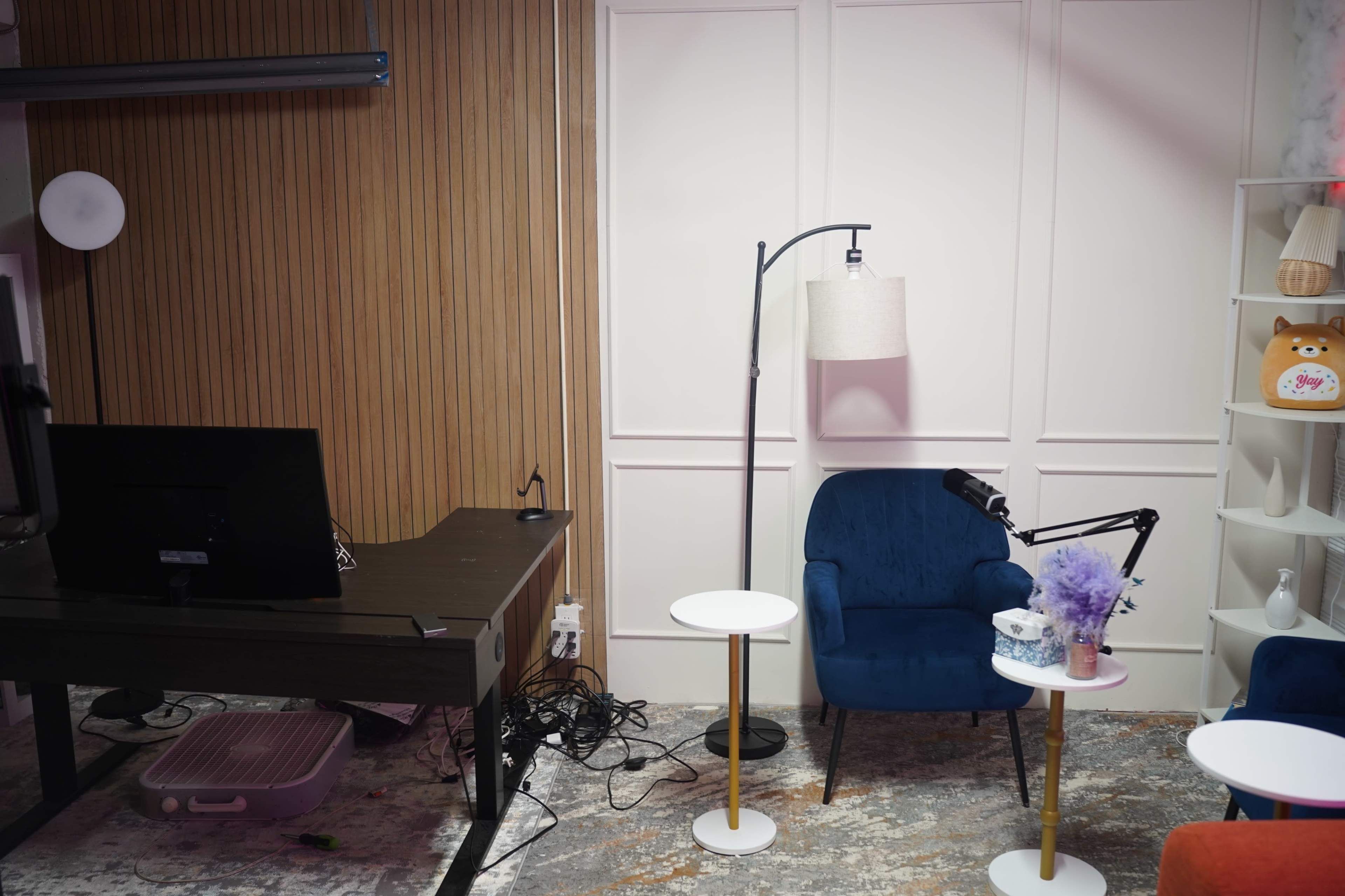 The image shows a cozy room with a blue armchair, a small white table, a desk with a computer monitor, and a floor lamp, set against a backdrop of wood paneling and white walls.