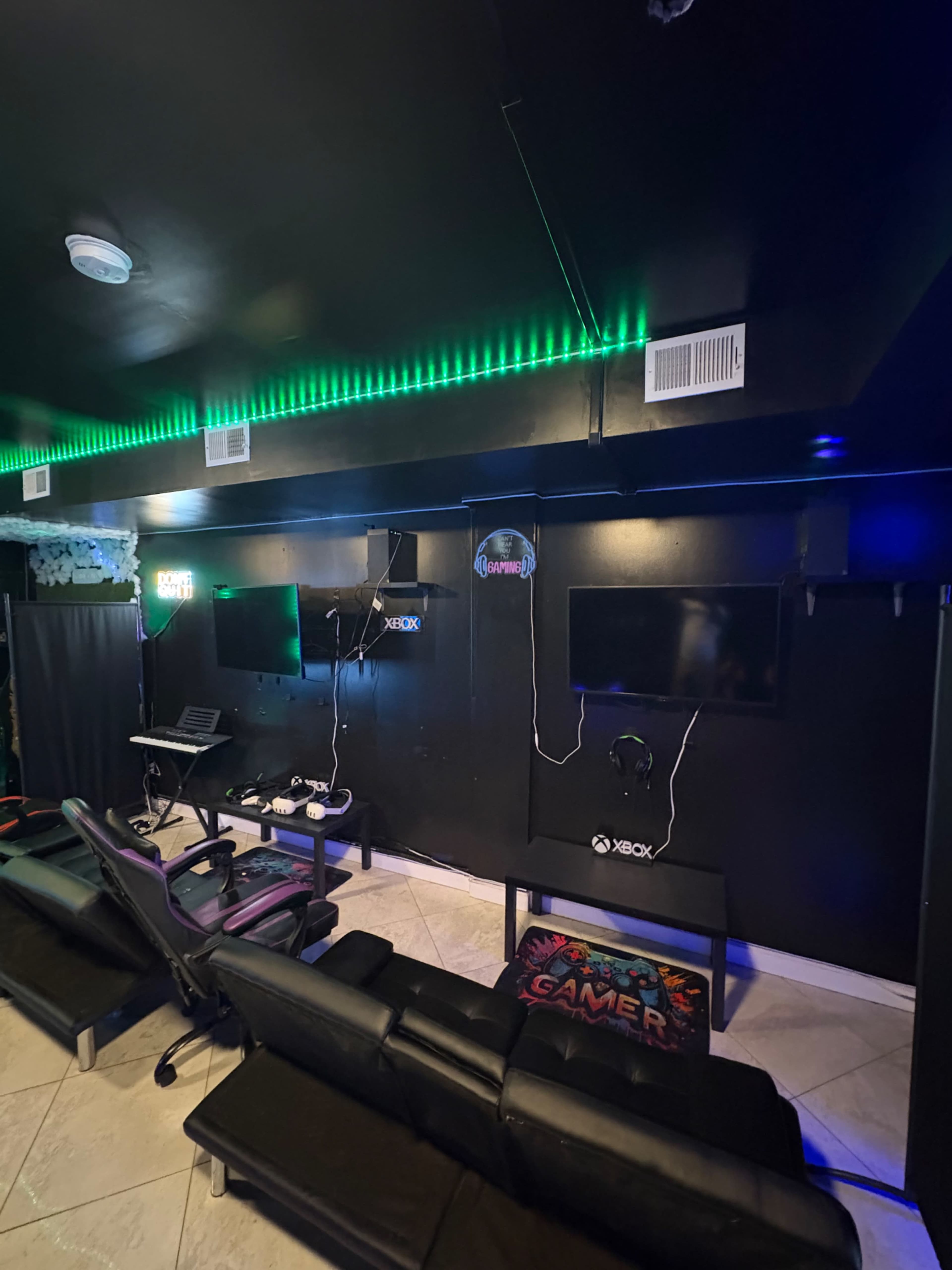 The image shows a gaming room with black walls, green LED lighting, two mounted screens, and a black sectional sofa.