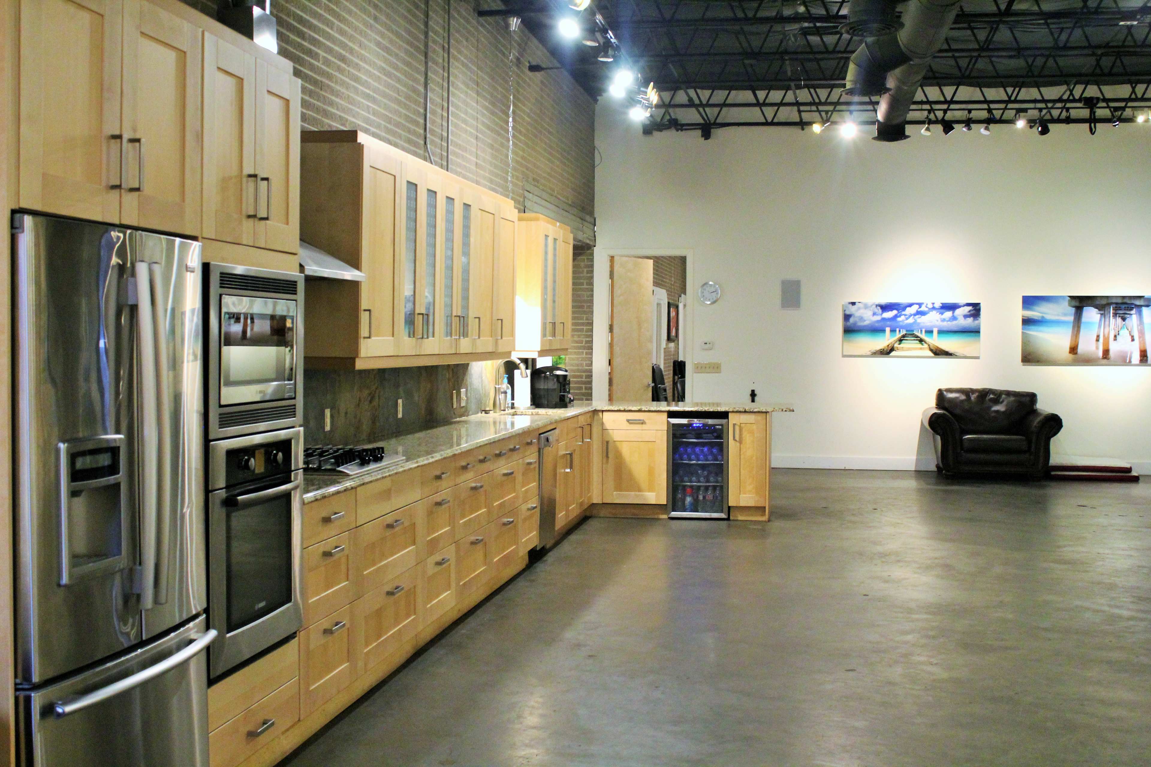 The image shows a modern kitchen with wooden cabinetry, stainless steel appliances, and a spacious layout, adjacent to a gallery-like area featuring two framed photographs and a leather sofa.