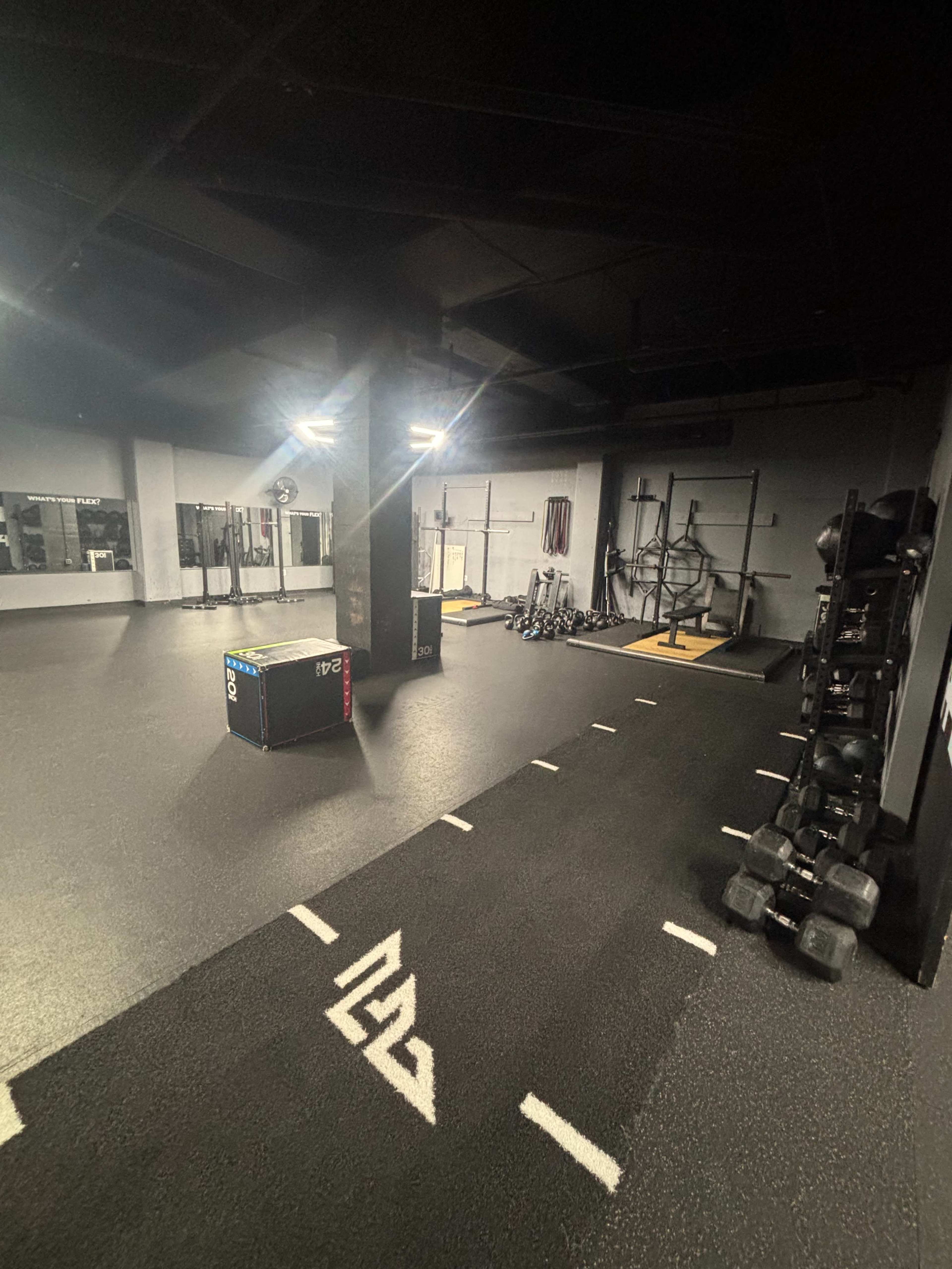 The image shows an indoor gym with workout equipment, including weights and a plyometric box, situated on a dark, rubberized floor.