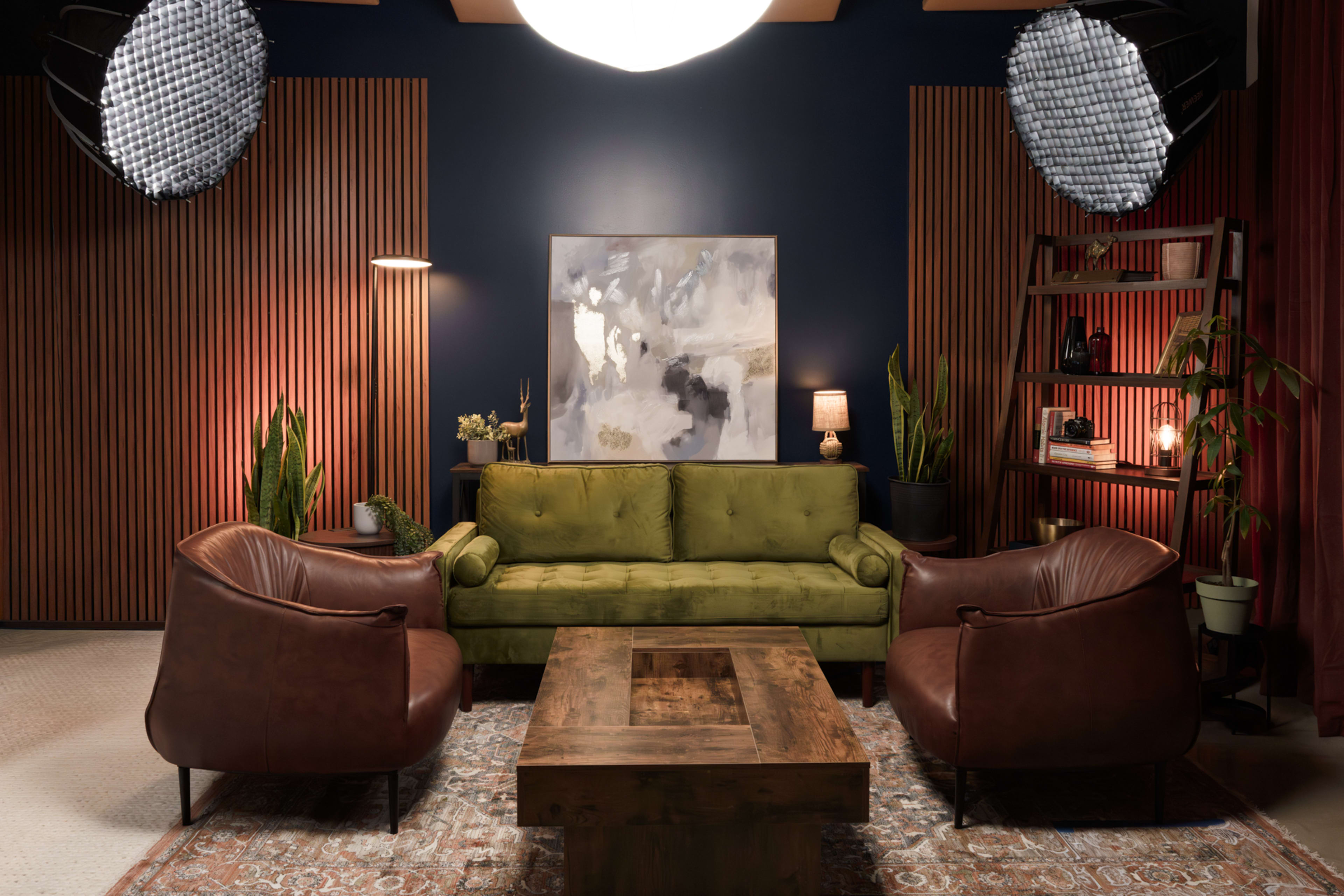 The image shows a stylish living room with a green velvet sofa, two brown leather chairs, a wooden coffee table, and artistic wall decor, all set against a backdrop of dark blue walls and wooden paneling.