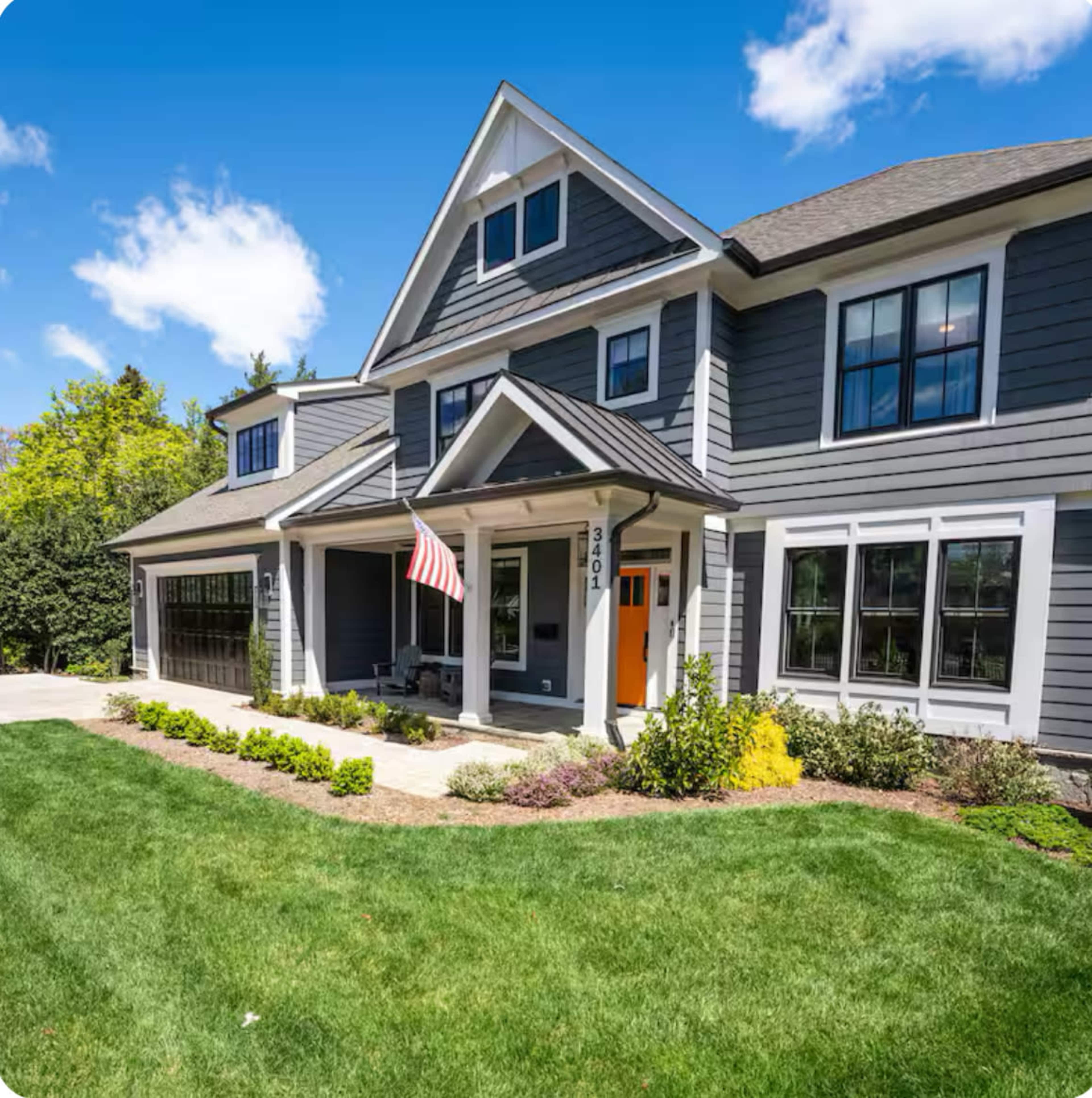 The image shows a two-story gray house with white trim, a front porch, and an American flag, surrounded by a well-maintained lawn and garden.