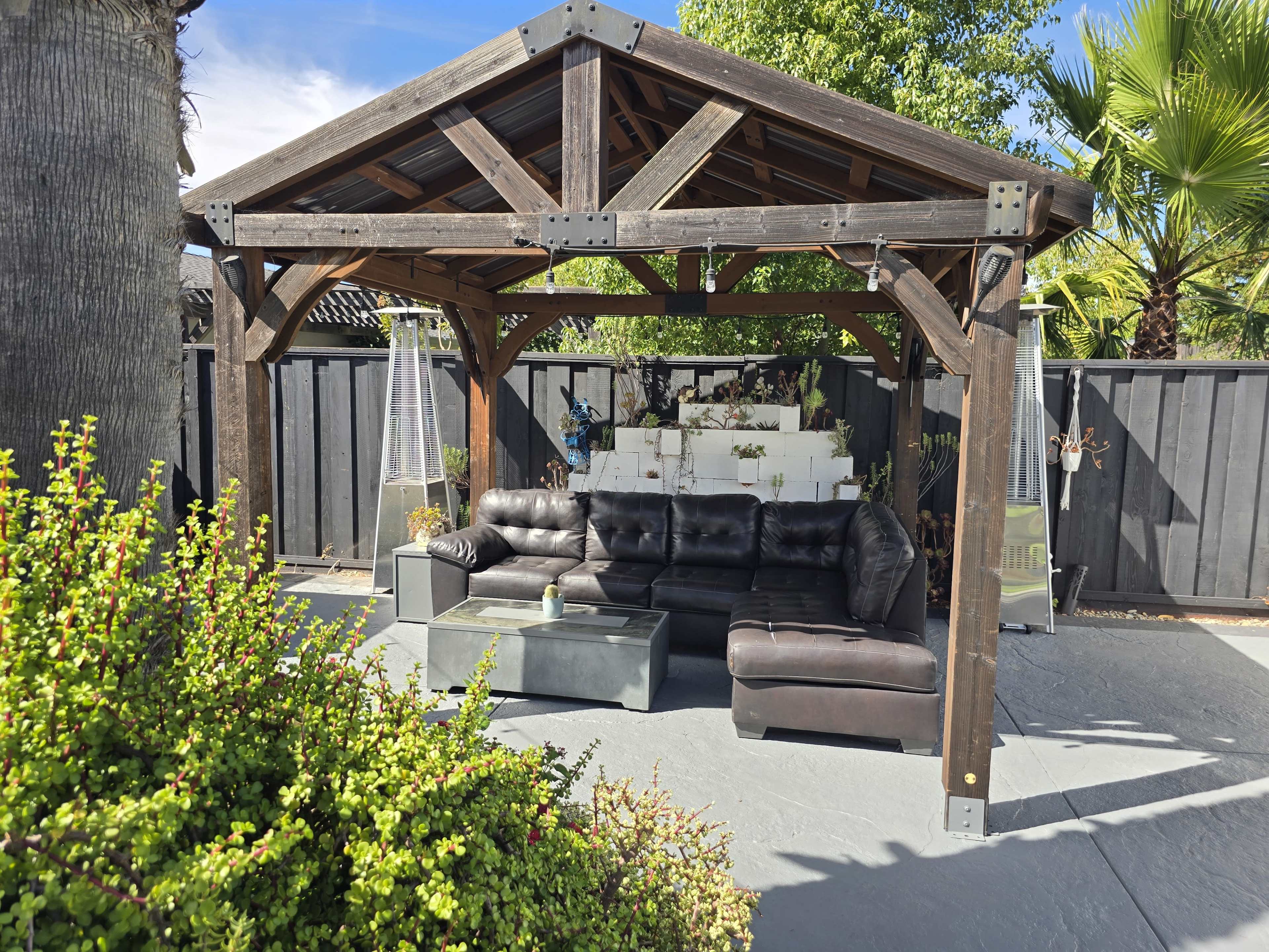 The image shows a wooden gazebo with a black sectional sofa and a glass coffee table, surrounded by greenery and a black fence.