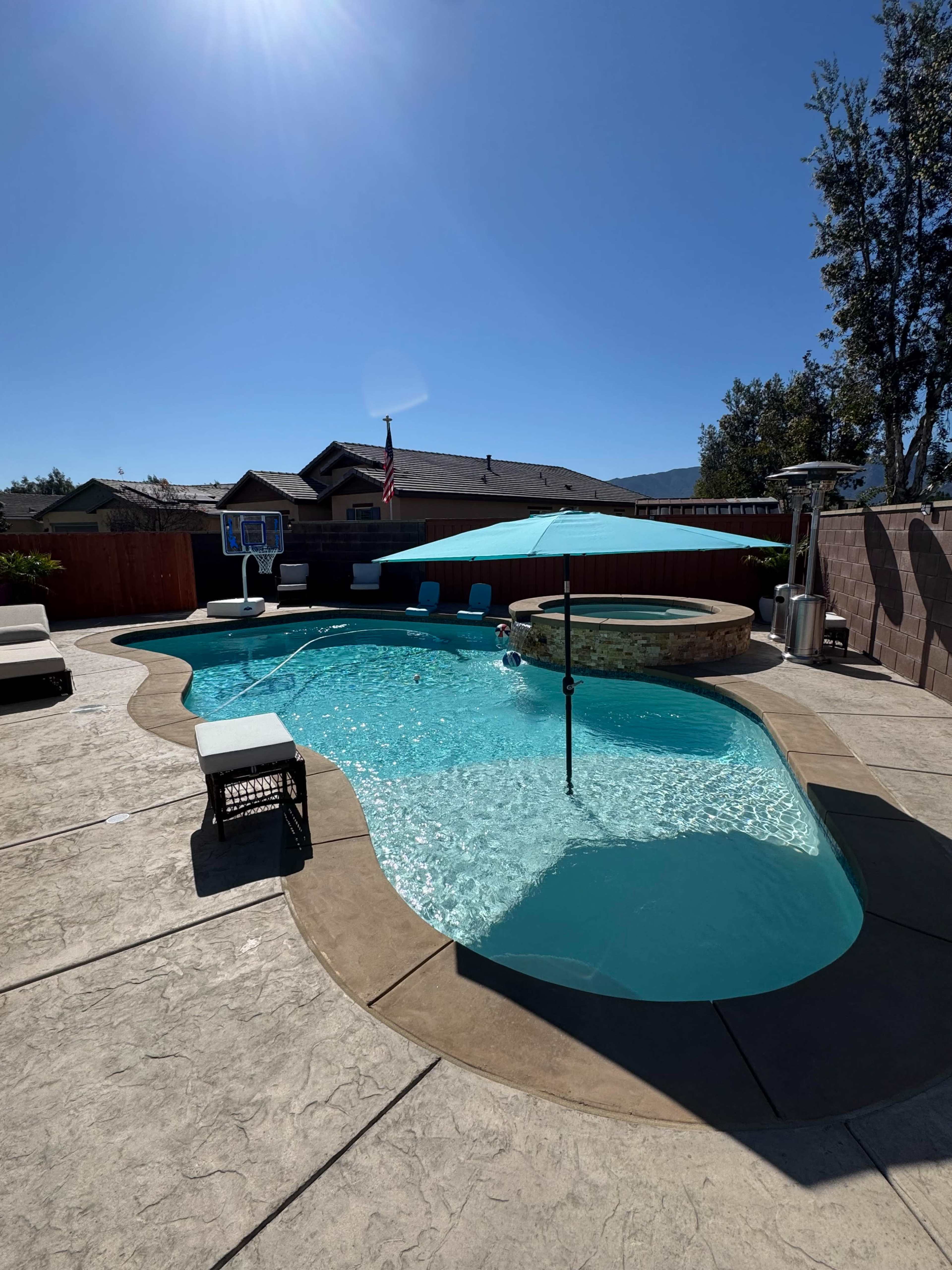 A curved swimming pool with a hot tub and patio furniture is set in a sunlit backyard.