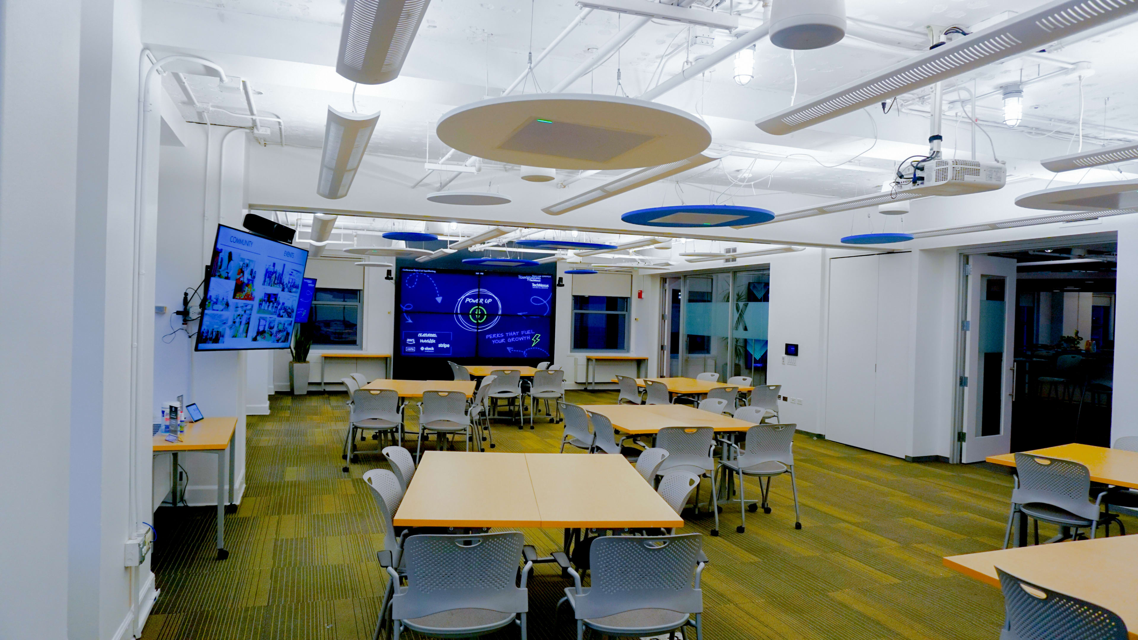 The image shows a modern classroom or meeting space with several tables and chairs, a large screen displaying information, and a bright, open layout with overhead lights.