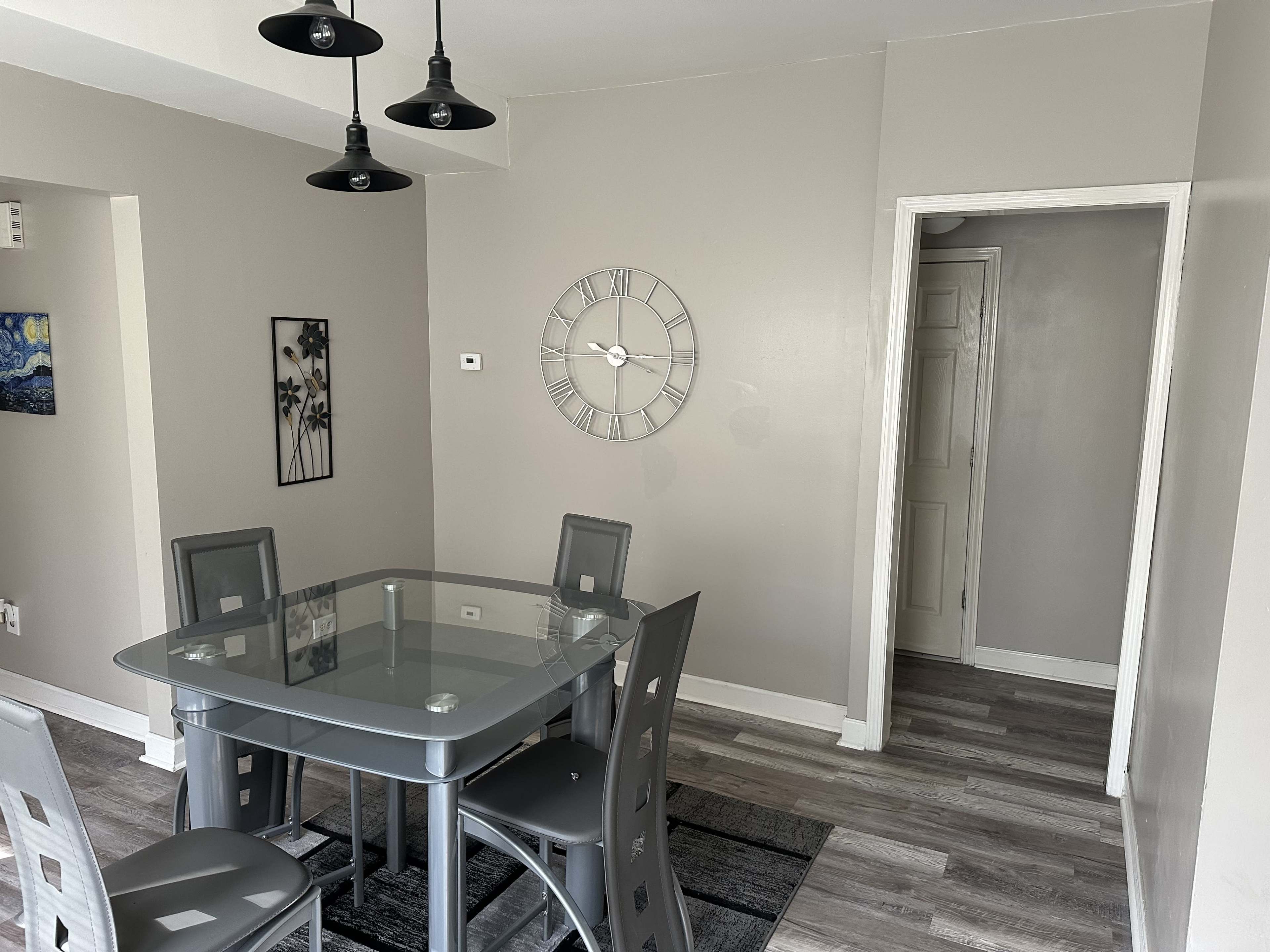 A glass-top dining table with gray chairs is positioned in a well-lit room featuring light gray walls and wooden flooring.