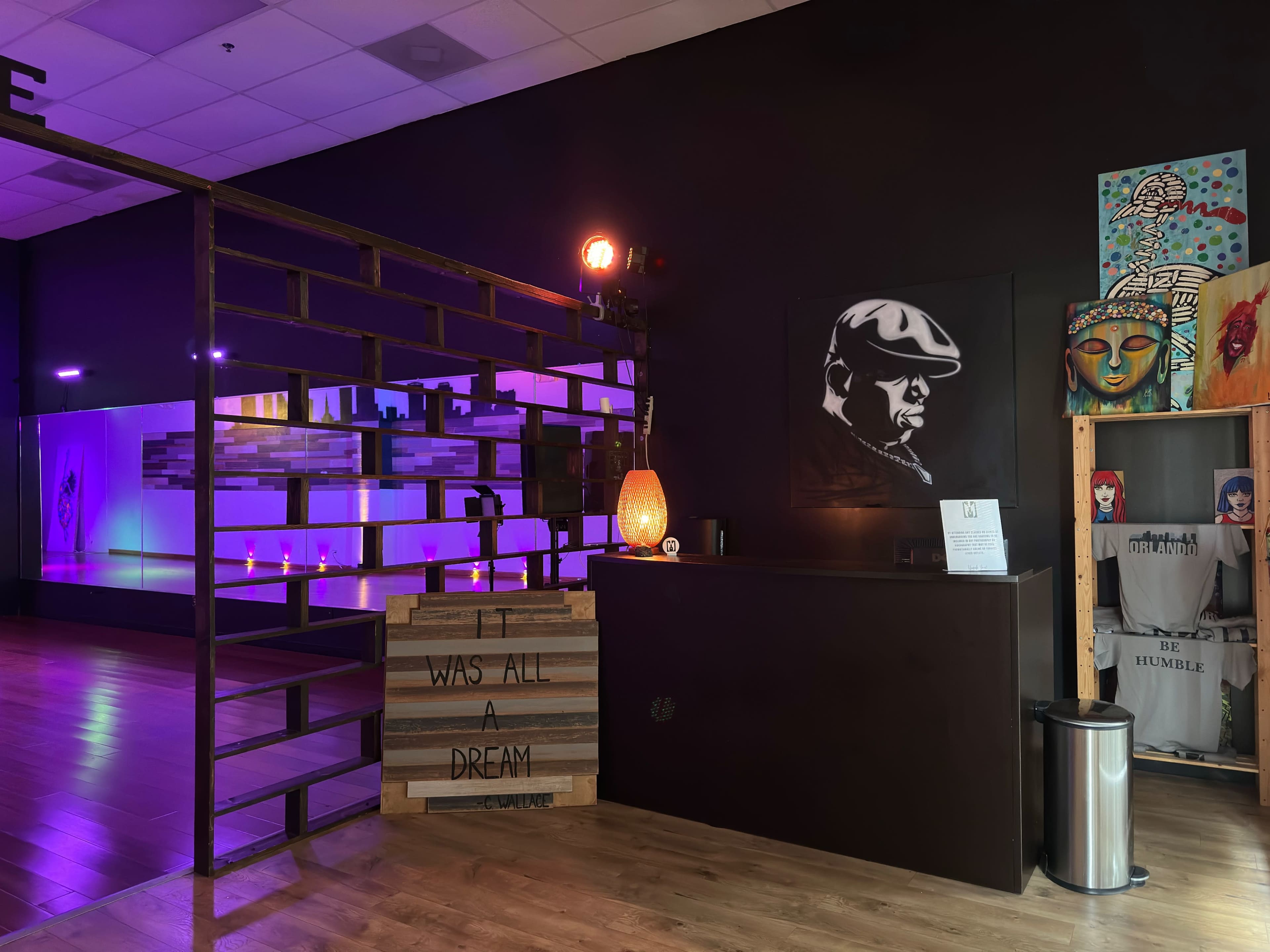 A dimly lit interior featuring a reception area with a wooden desk, artwork on the walls, and a staircase made of metal and wood.