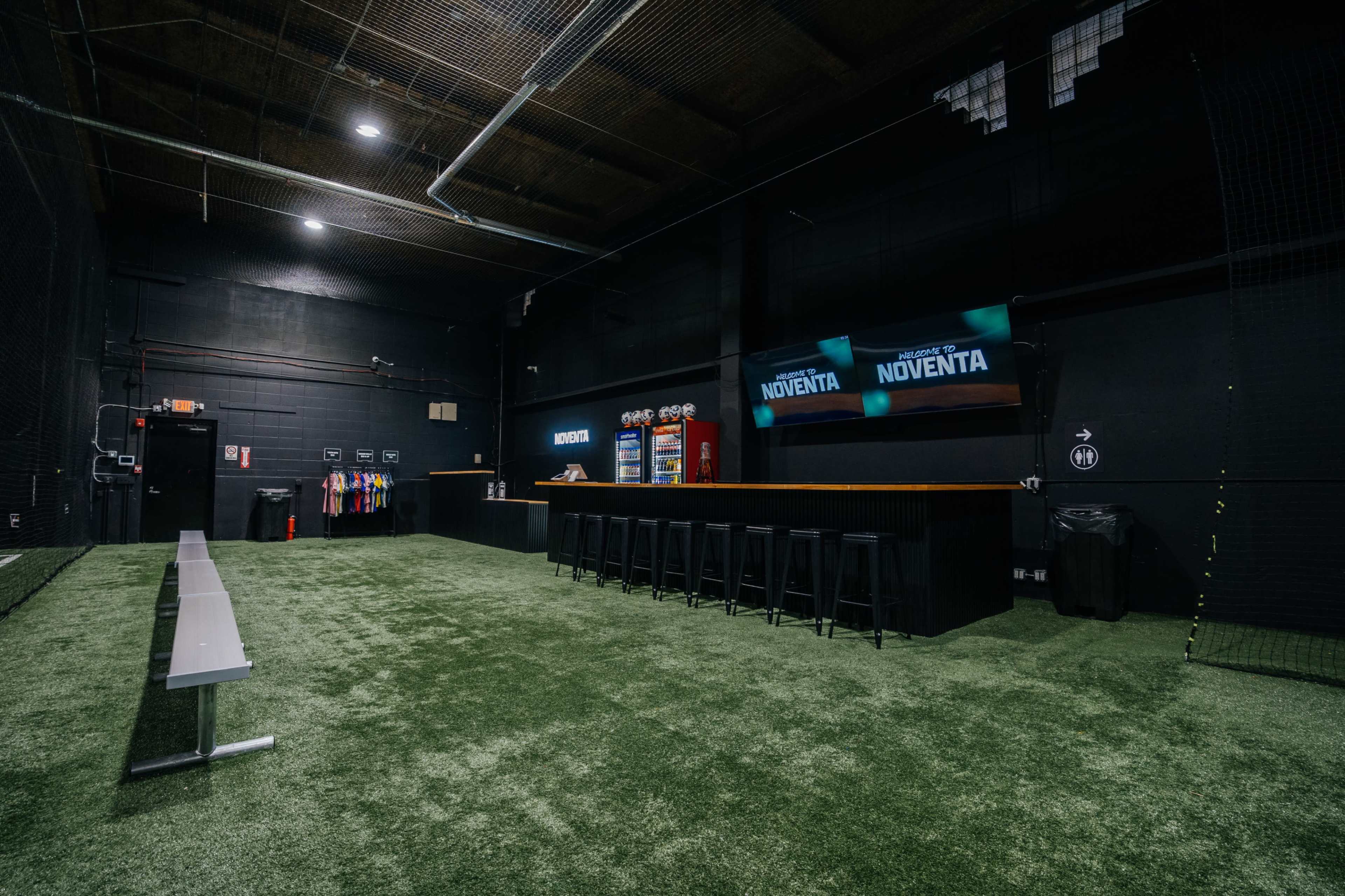 The image shows an indoor sports training facility featuring artificial turf, a bar-like seating area, and two large screens on the wall.