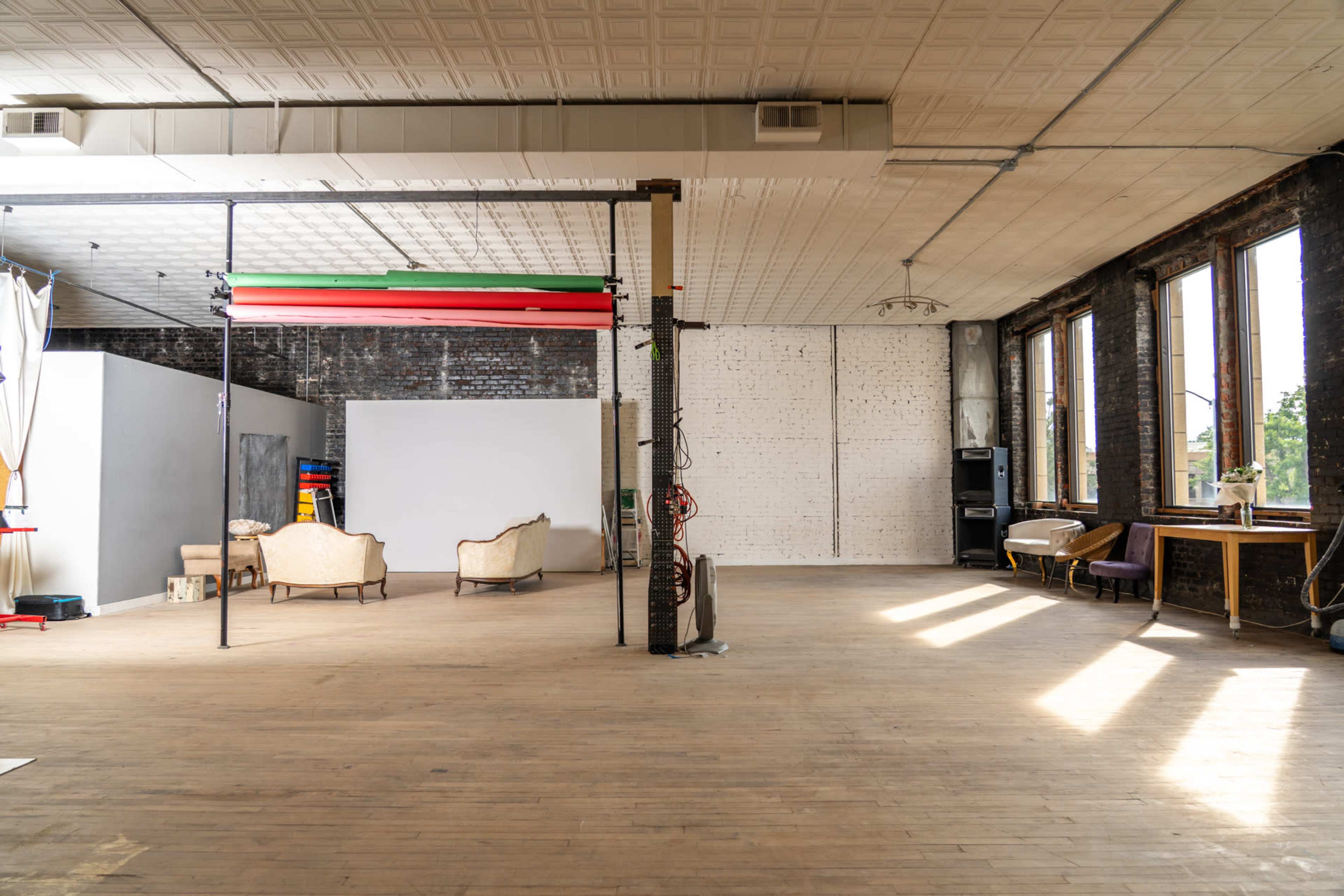 The image shows a spacious, empty studio with wooden floors, large windows, and simple furniture arranged on one side.