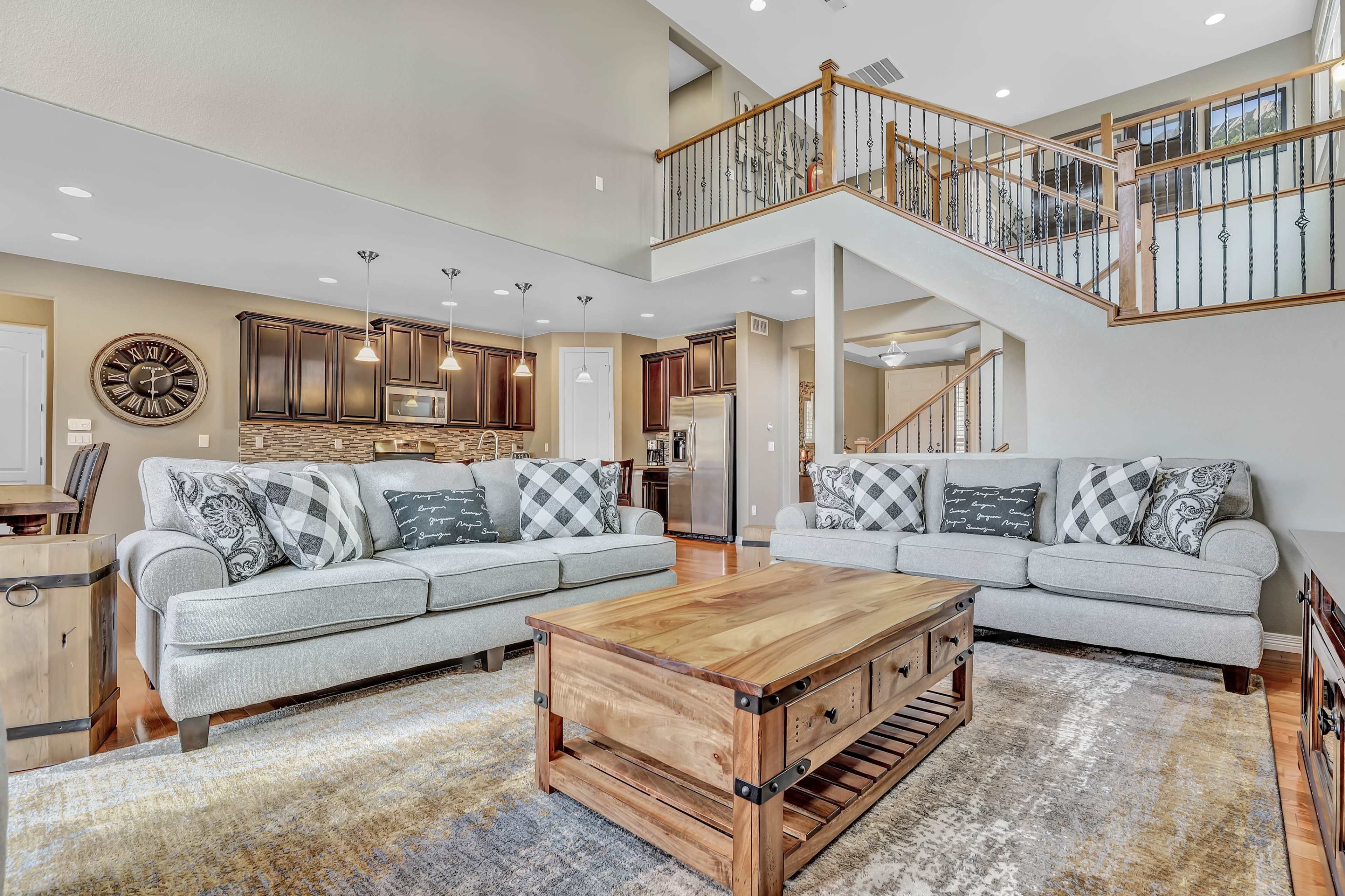The image shows a modern living room with two light gray sofas, a wooden coffee table, and an open layout connecting to a kitchen and dining area.