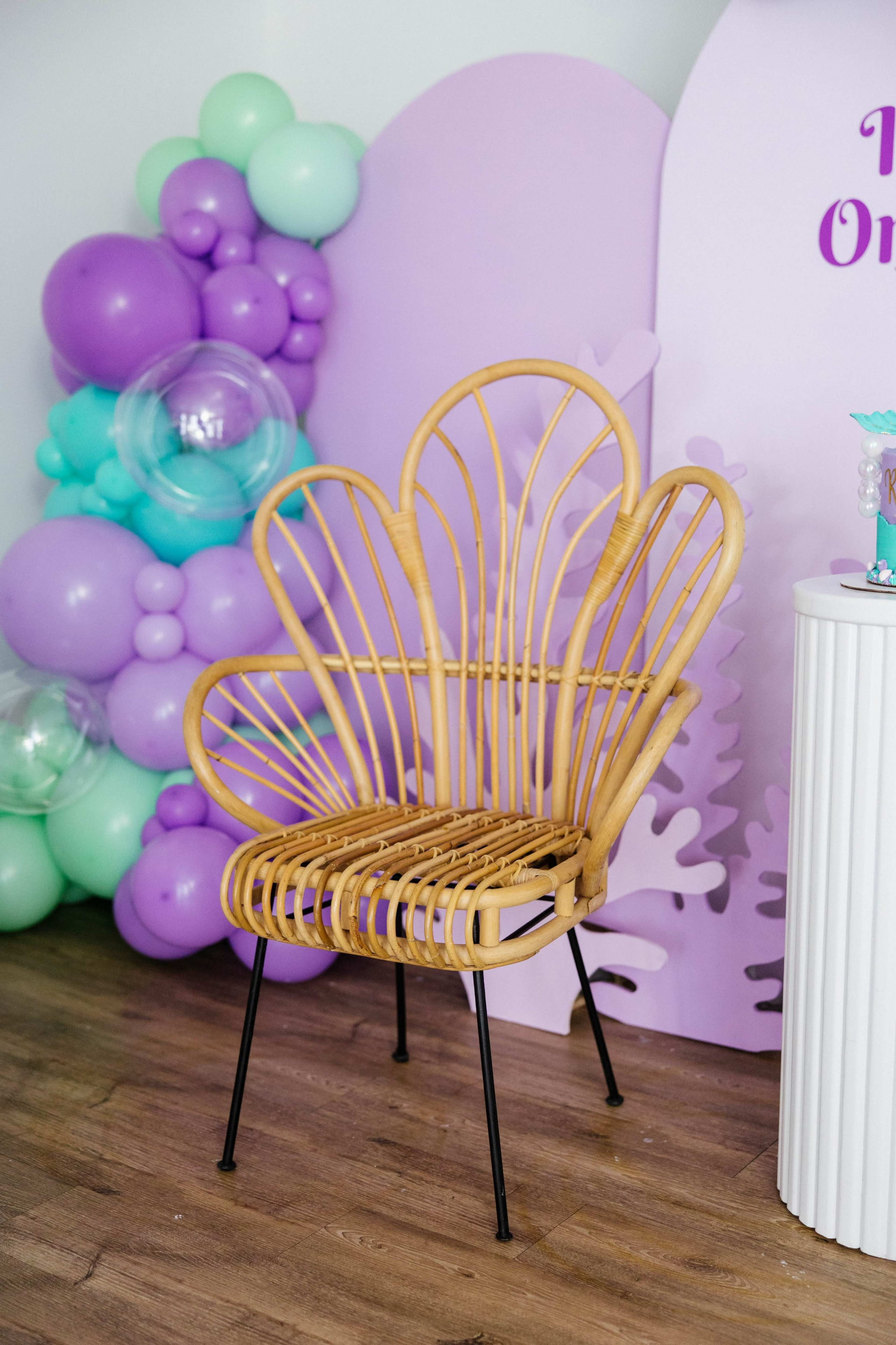 A rattan chair with a shell-shaped backrest positioned beside a cluster of pastel-colored balloons and a decorative backdrop.