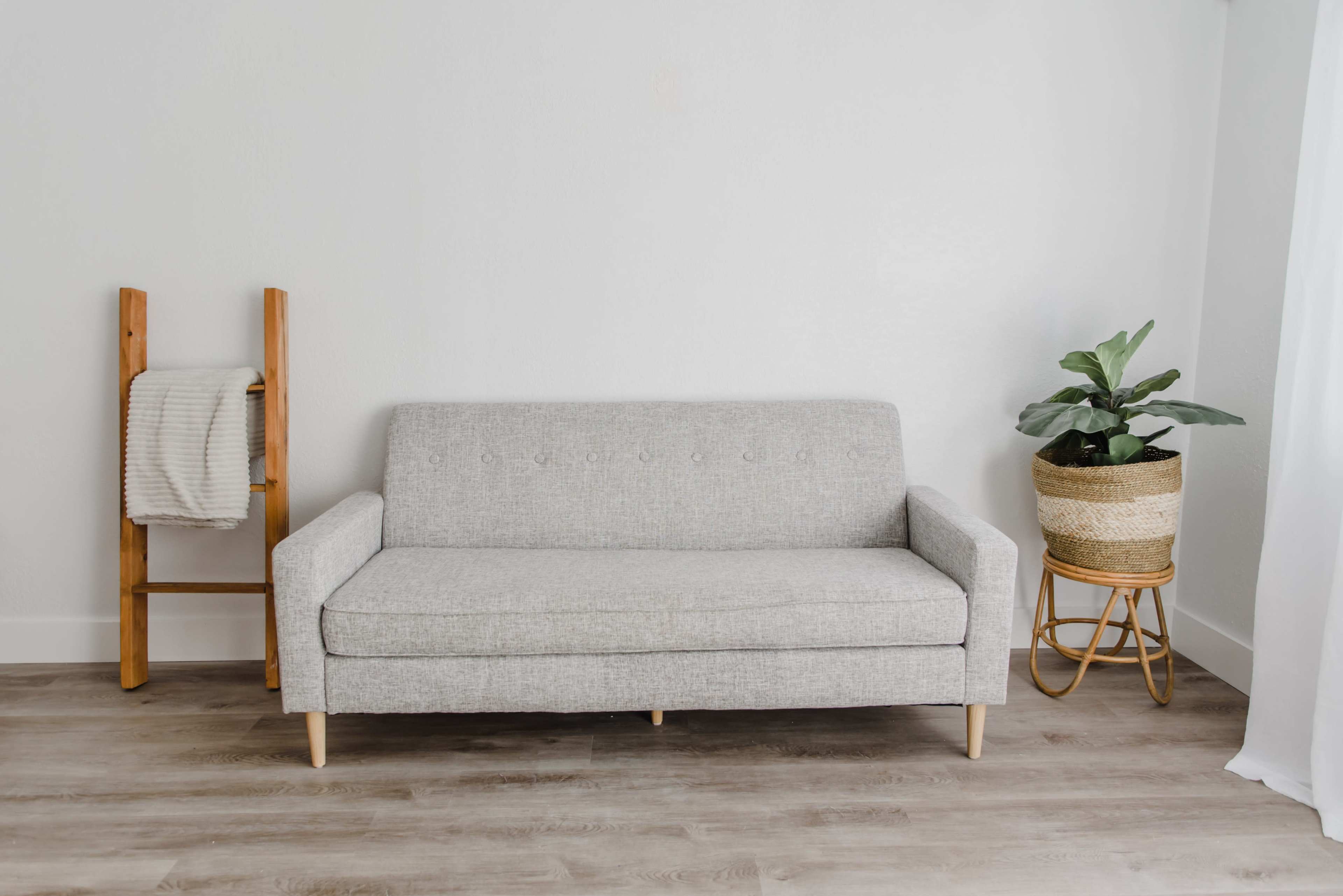 A gray couch is positioned against a white wall, accompanied by a wooden ladder holding a blanket and a potted plant in a woven basket.