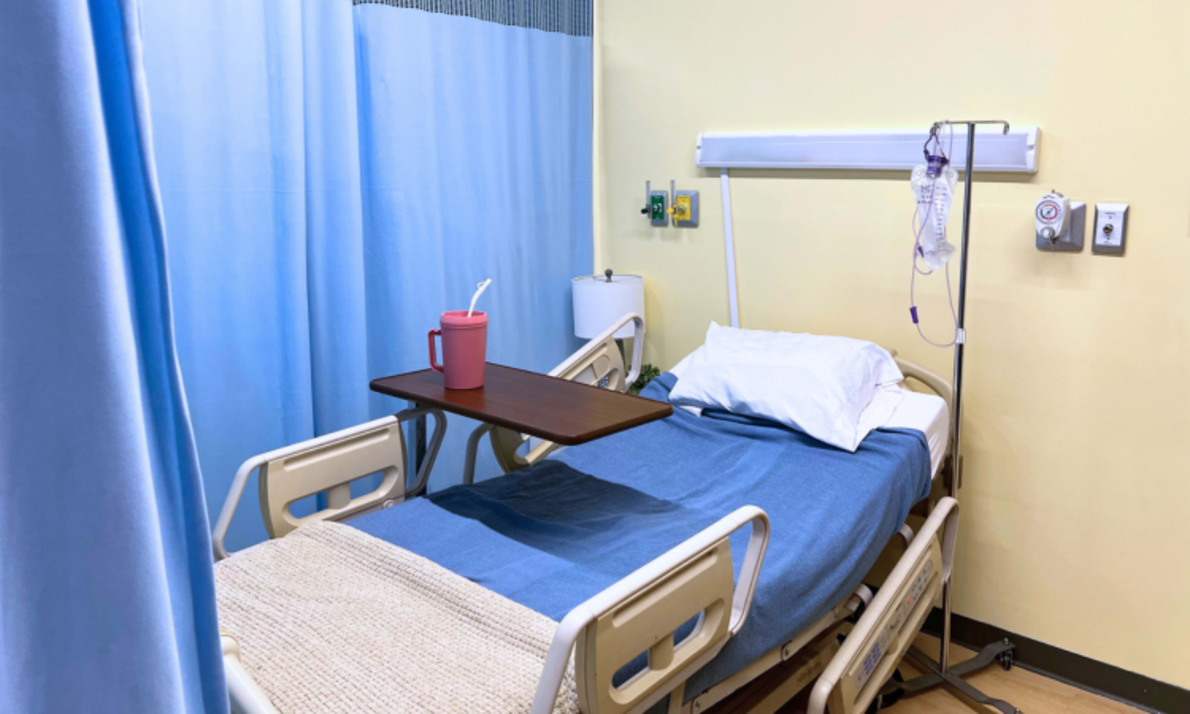 The image shows an empty hospital room with a patient bed, a side table, and a beverage cup on the table.