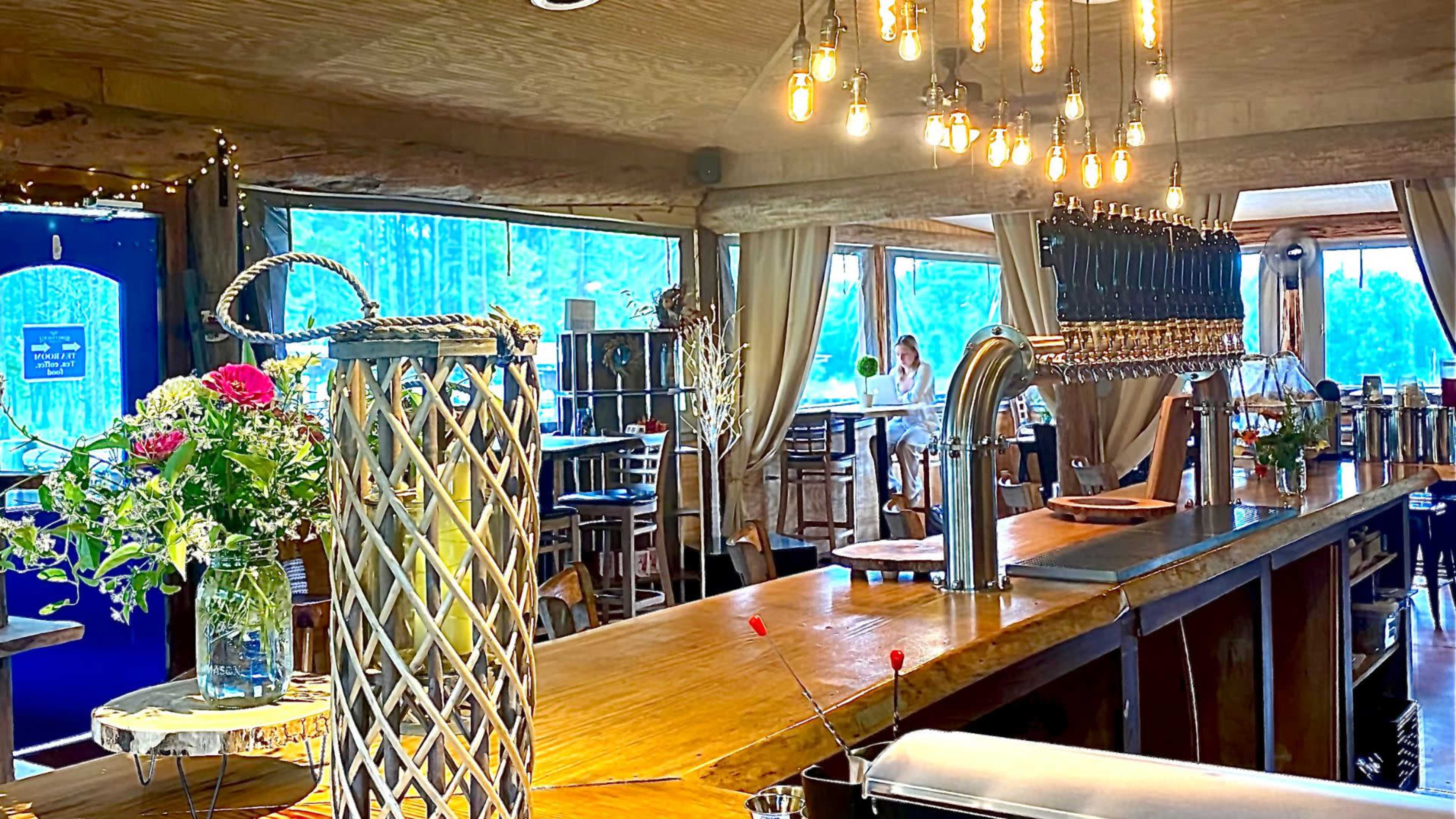 The interior of a bar featuring a wooden counter with several beer taps, surrounded by tables and large windows letting in natural light.