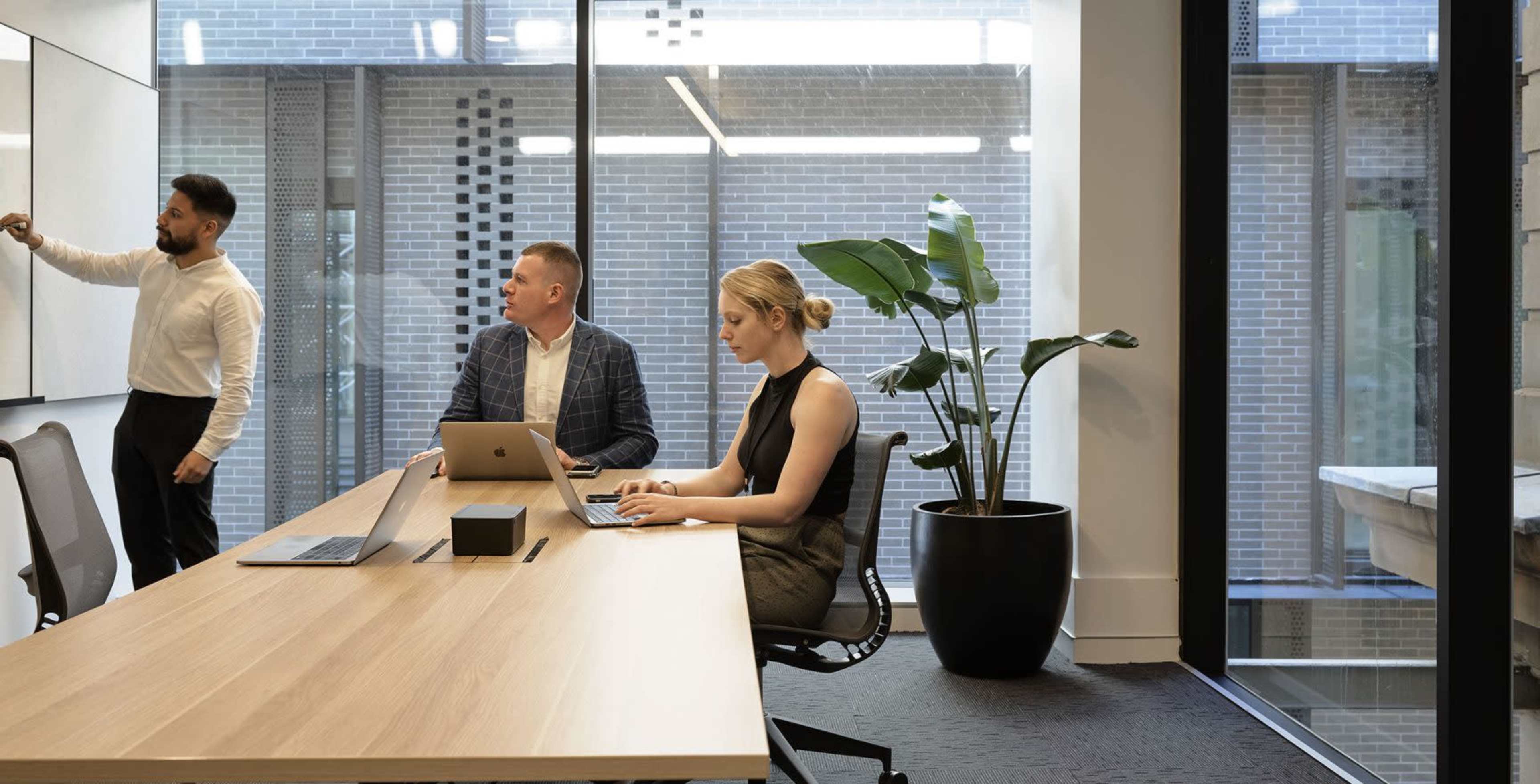 A man writes on a whiteboard while two colleagues sit at a conference table, one working on a laptop and the other observing.