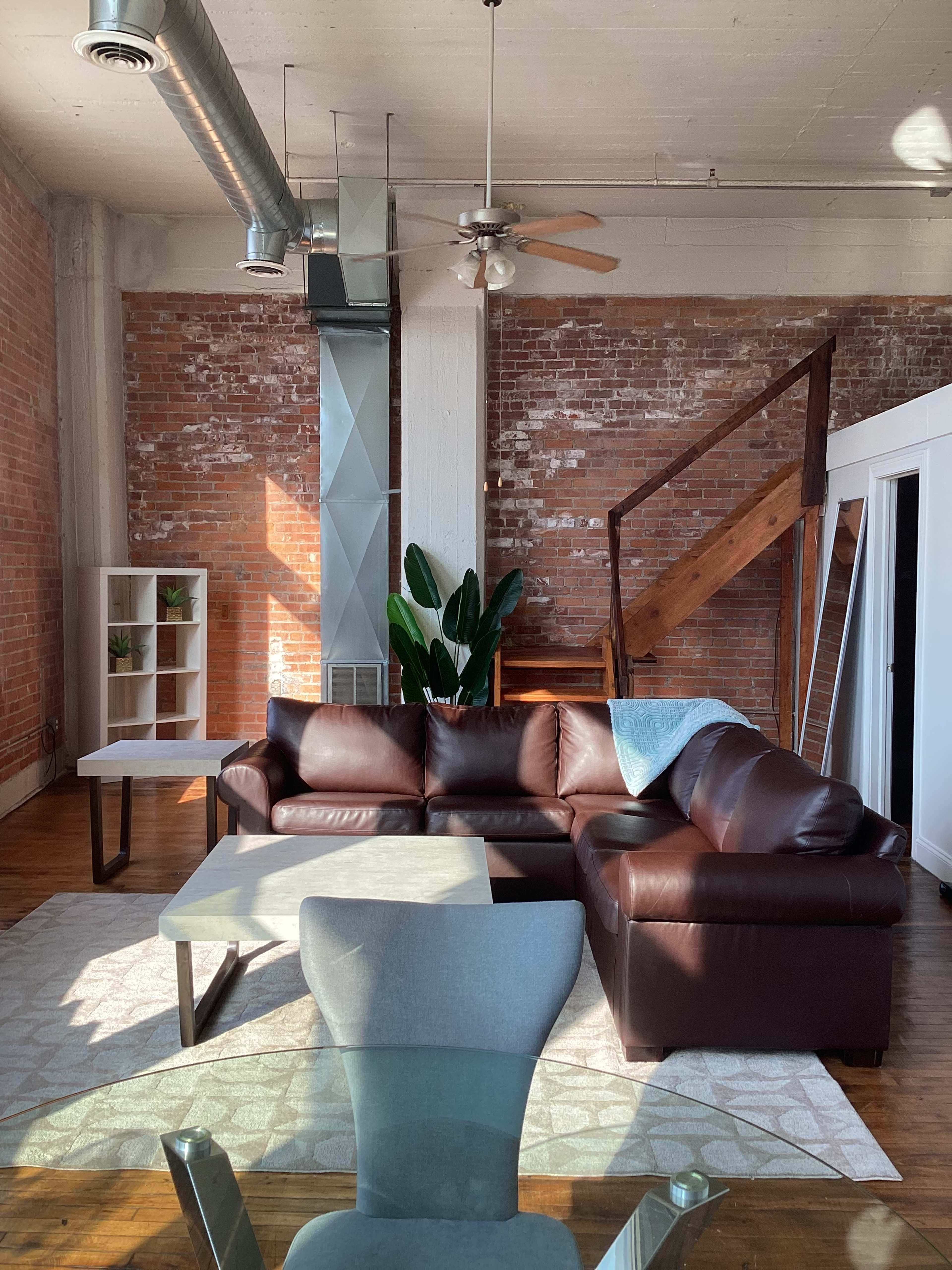 The image shows a modern living room with a brown sectional sofa, a glass coffee table, and a small dining area, set against exposed brick walls and featuring a staircase in the background.