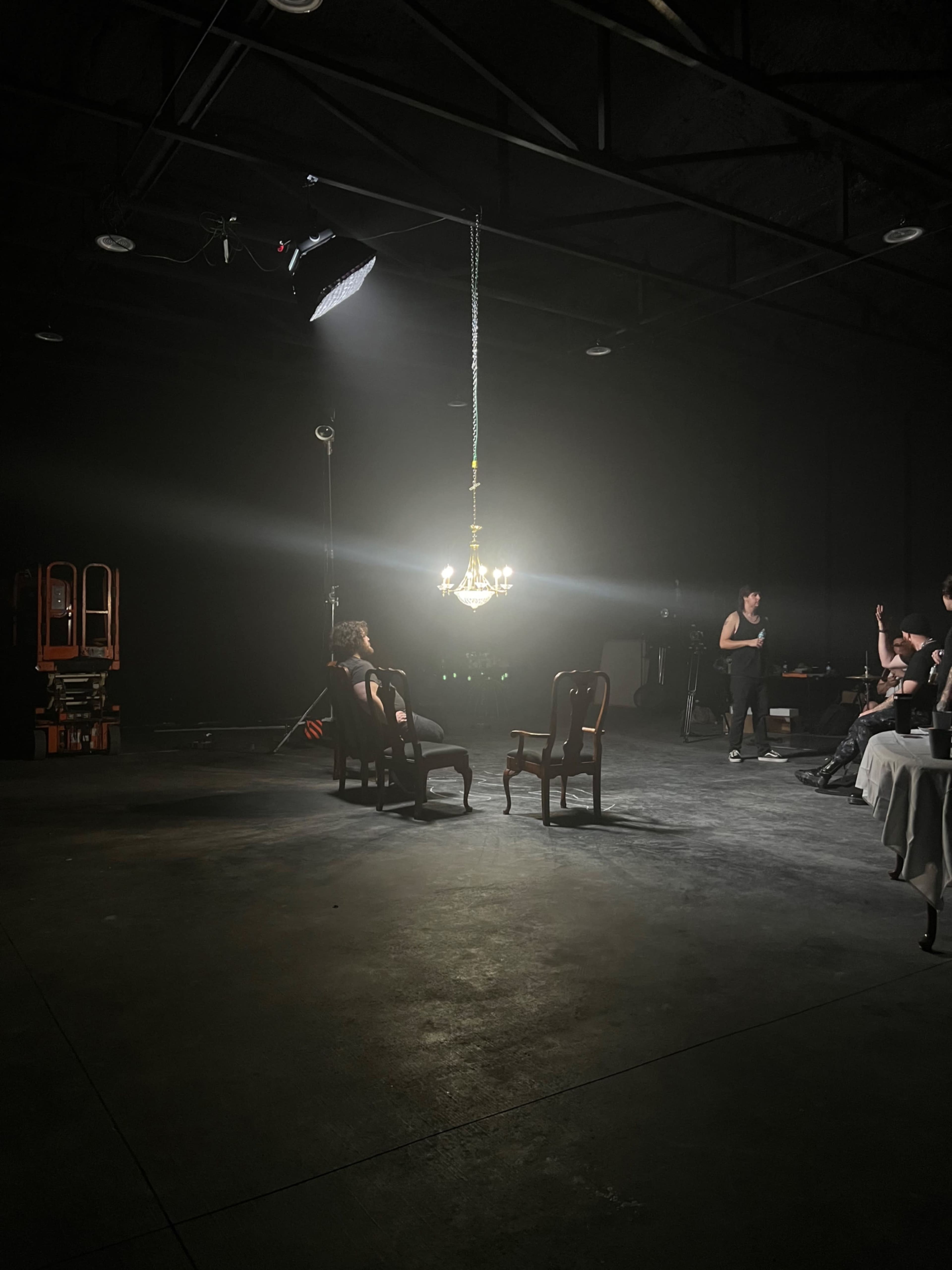 A dimly lit studio space features two chairs illuminated by a chandelier, with crew members and equipment in the background.
