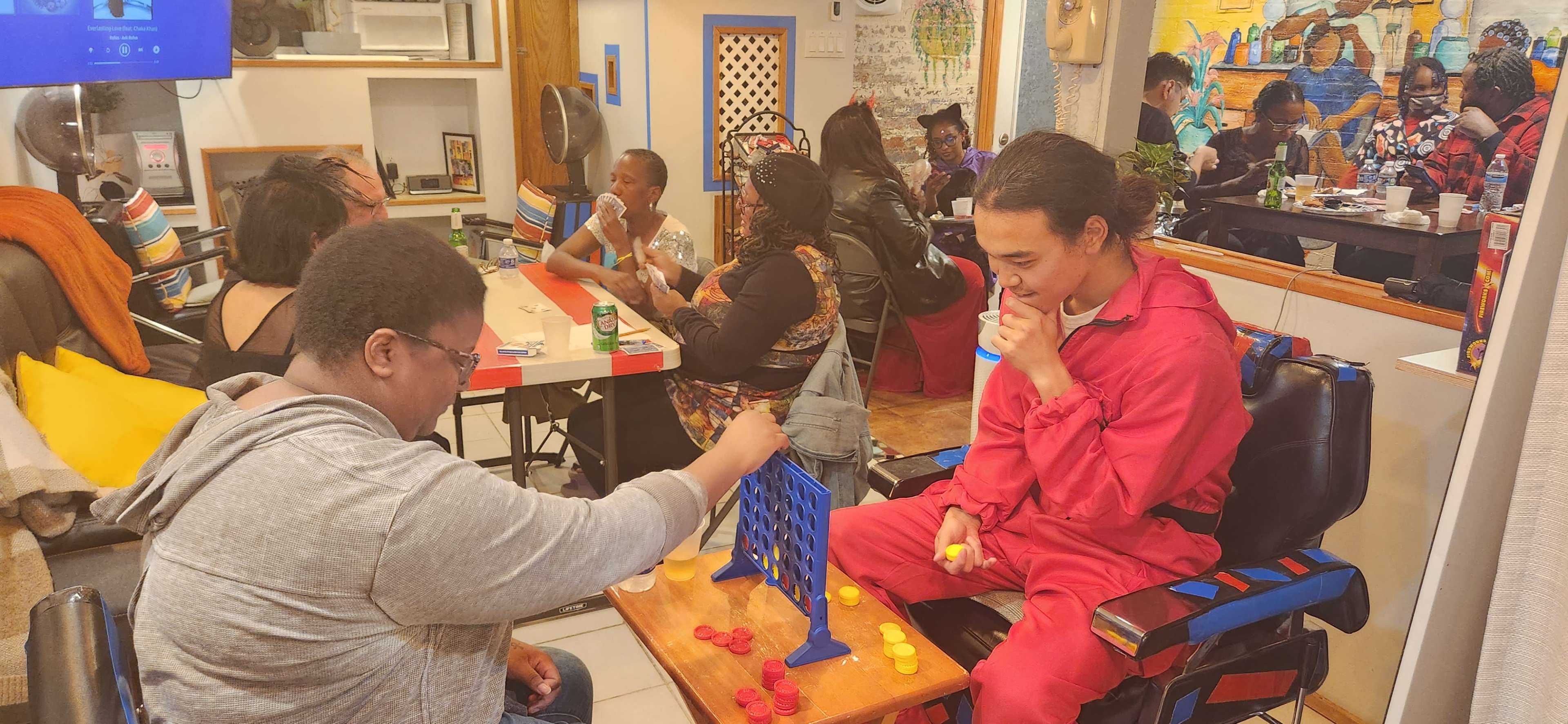 A group of people socializes in a lively café while two individuals play a game of Connect Four at a table in the foreground.