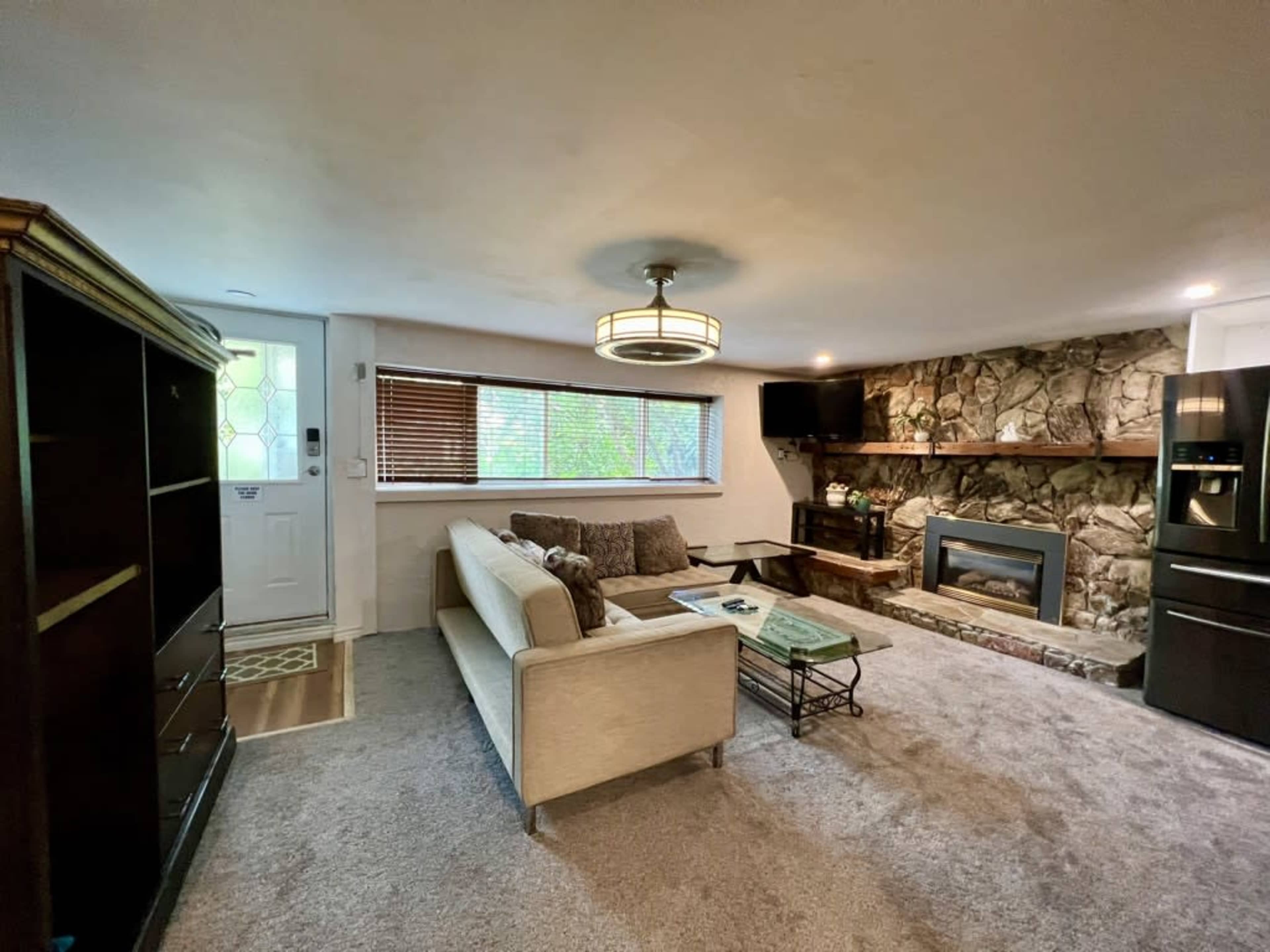 The living room features a beige sofa, a stone fireplace, and a large window with wooden blinds.