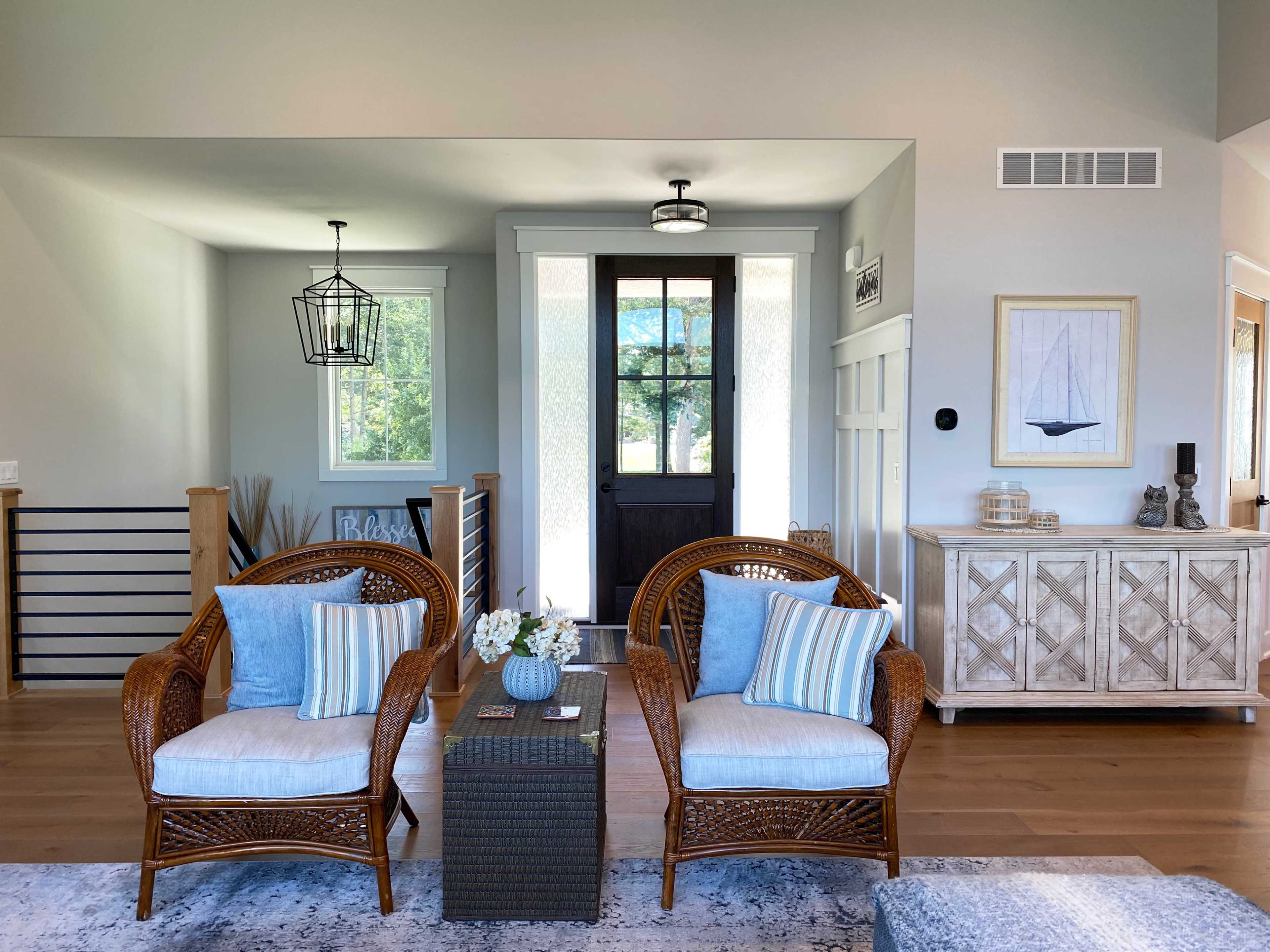 A bright living room with two wicker chairs, a coffee table, and a decorative sideboard, all arranged around a dark front door.